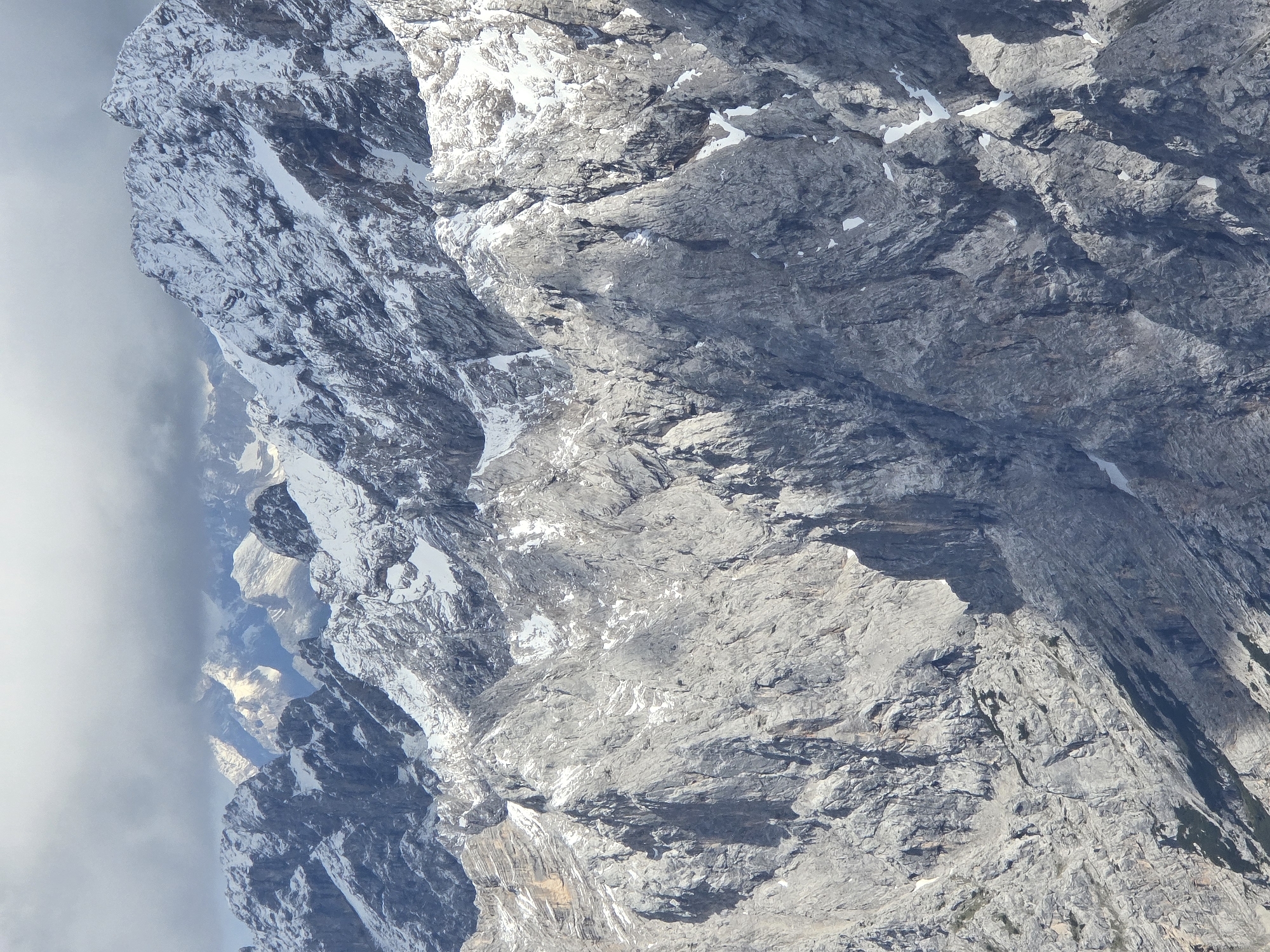 The image depicts a close-up, telephoto view of a rugged, rocky mountain range under a cloudy sky. The foreground is dominated by steep, grey, and textured rock faces, showing numerous cracks, crevices, and ledges. Patches of snow are scattered across these rocks, particularly in shadowed areas and on higher elevations. In the background, the mountain peaks are more heavily capped with snow and appear sharper and more defined, though some are obscured by dark, heavy clouds. Sunlight intermittently breaks through the clouds, illuminating sections of the snow-covered peaks with a bright, almost ethereal glow. The overall impression is one of a harsh, remote, and imposing alpine environment. This scene is likely a view of the Zugspitze massif, the highest peak in Germany, located near Garmisch-Partenkirchen, Bavaria.