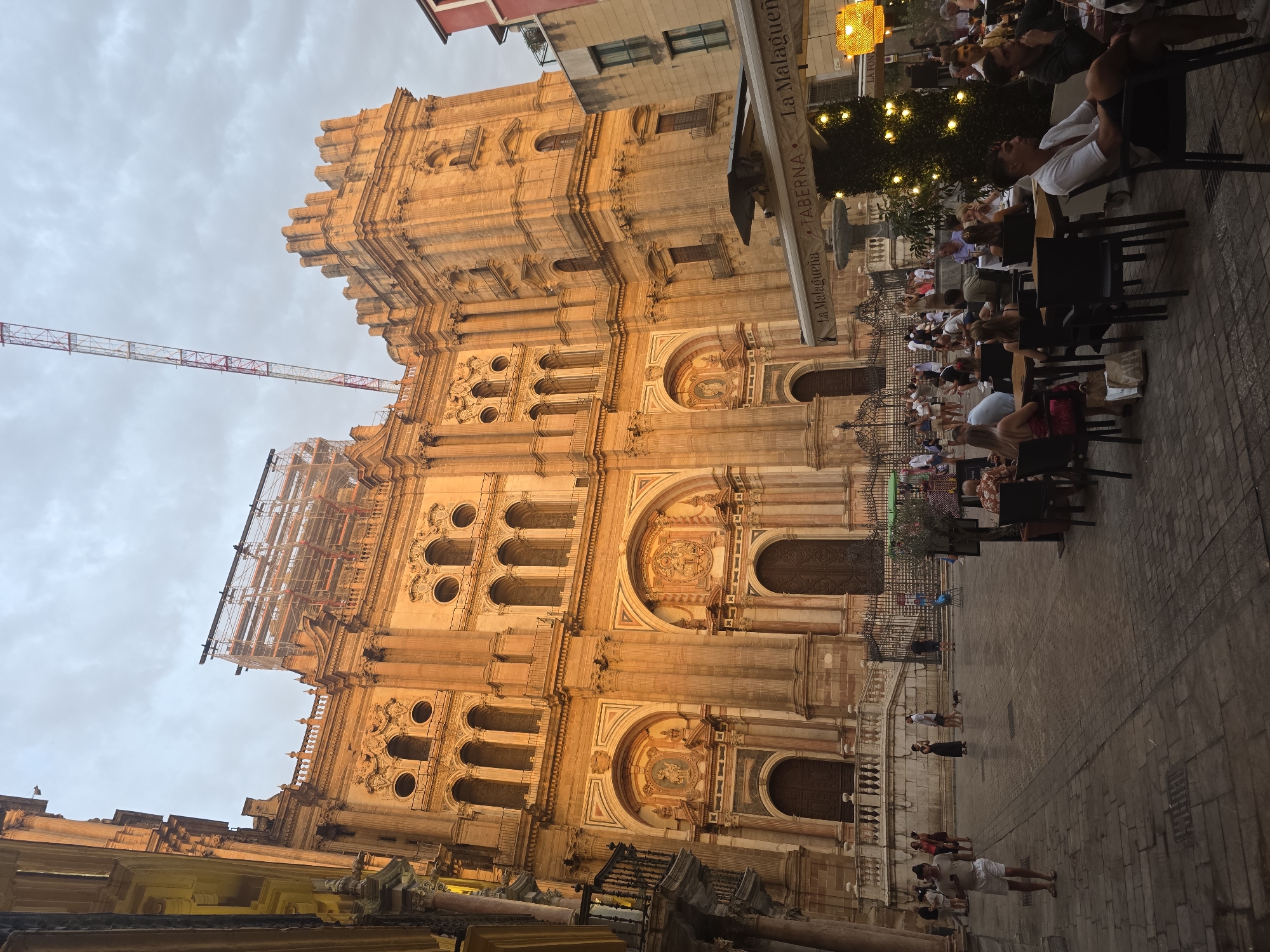 The image depicts the Malaga Cathedral in Malaga, Spain, with a large crane and scaffolding on its upper section. The cathedral is a grand, ornate structure made of golden-hued stone. In the foreground, a plaza is filled with outdoor seating for a cafe or restaurant. People are seen sitting at tables, dining, and socializing. The plaza is paved with cobblestones, and the scene is illuminated by the warm glow of the setting sun or artificial lighting. To the right of the cathedral, other buildings are visible, including a taller, modern building with balconies. A Christmas tree decorated with lights stands to the right, suggesting the image may have been taken during the holiday season.
