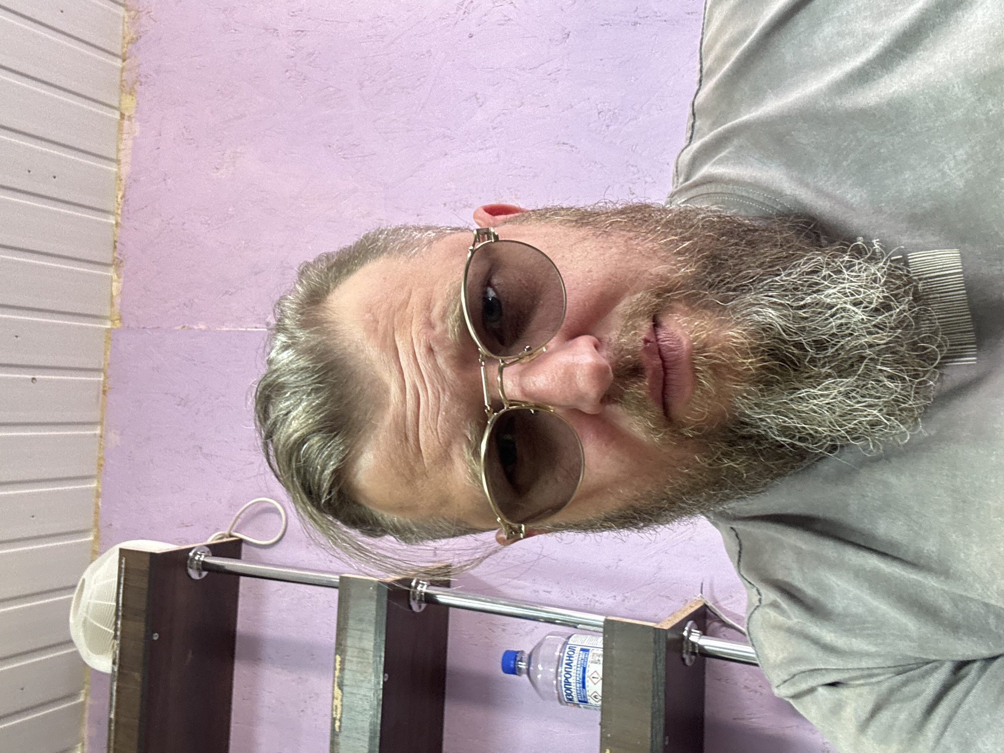 A man with graying hair and a full beard wearing sunglasses and a grey t-shirt is pictured in a close-up, head-and-shoulders shot. He is looking directly at the camera. Behind him, a shelf unit with multiple shelves is visible. On one of the shelves sits a clear plastic bottle with a blue cap and a white label that reads "ИЗОПРОПАНОЛ". The wall behind the shelf unit is painted a light purple color. The ceiling above is made of white paneling. The environment appears to be indoors. The location is identified as Schokino, Russia.
