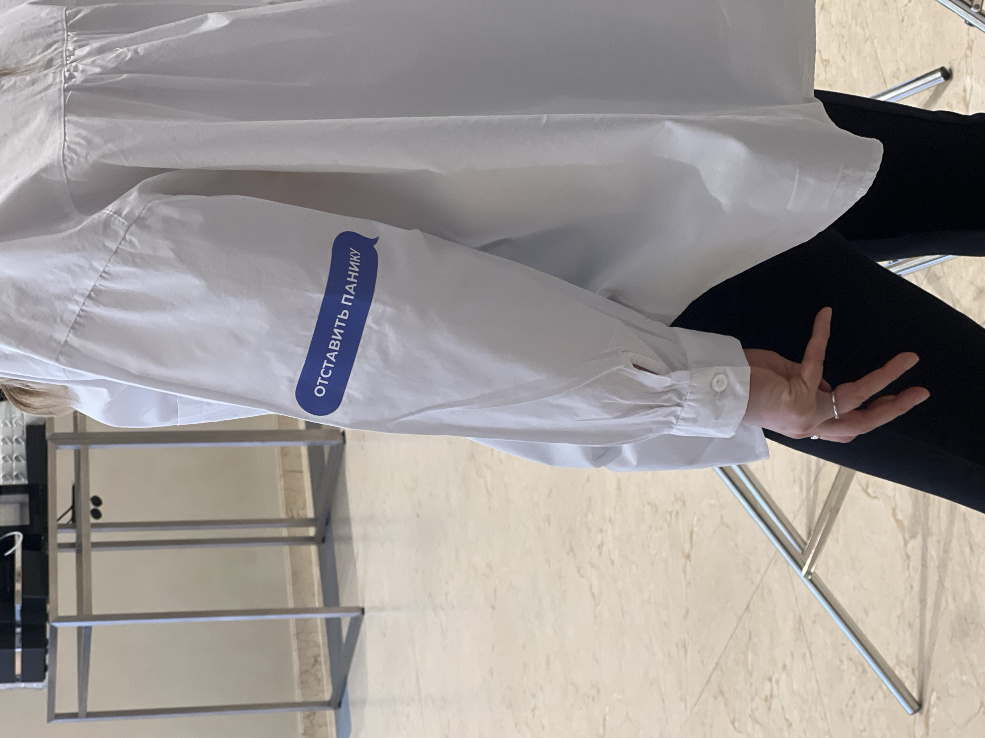 A person wearing a white shirt with a blue inscription in Russian "Отставить панику!", which translates to "Discard the panic!", is depicted. The shirt appears to be a long-sleeved, button-down style. The person also wears black pants. A metal stand is visible in the background. The location is in Moscow, Russia.