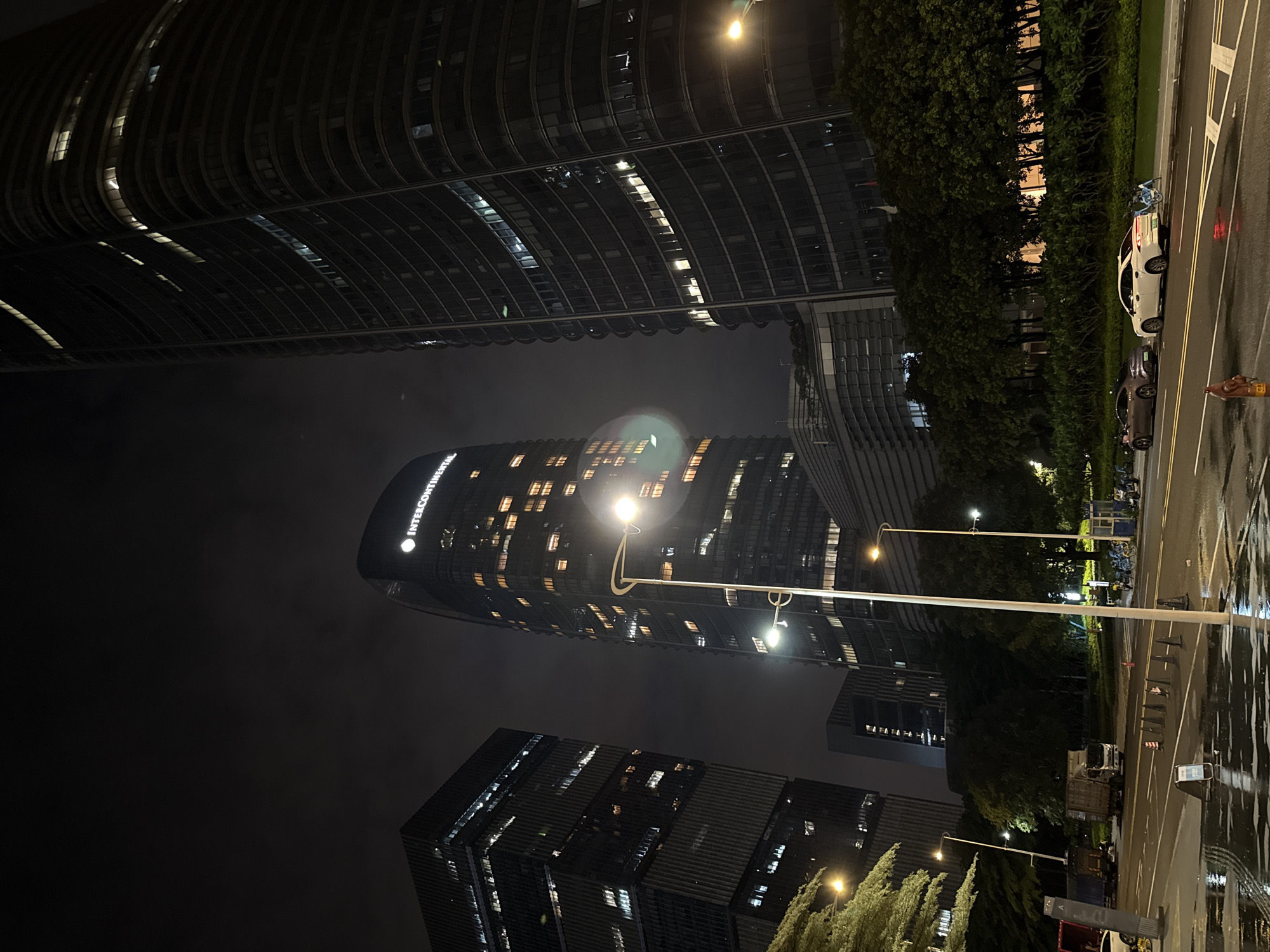 The image depicts a nighttime street scene in Haizhu District, China, with modern skyscrapers dominating the background. Two prominent buildings are visible: on the left, a rectangular skyscraper with numerous lit windows, and on the right, a curved skyscraper with the "INTERCONTINENTAL" logo and a circular emblem illuminated near its peak. Both buildings are dark-colored and reflect light from within.
In the foreground, a wet street reflects the ambient light from street lamps and buildings, suggesting recent rain. Several streetlights on tall poles are present, some illuminating the road and sidewalk. Parked along the side of the road are two cars, a dark-colored coupe and a white sedan. Several bicycles are also visible, some parked and some possibly in a designated docking area.
Further down the street, several large trucks are parked, one appearing to be a delivery truck. A line of traffic cones is placed on the road. Trees and dense shrubbery line the area between the road and the buildings, adding a natural element to the urban landscape. The overall atmosphere is that of a well-lit, developed urban area at night.