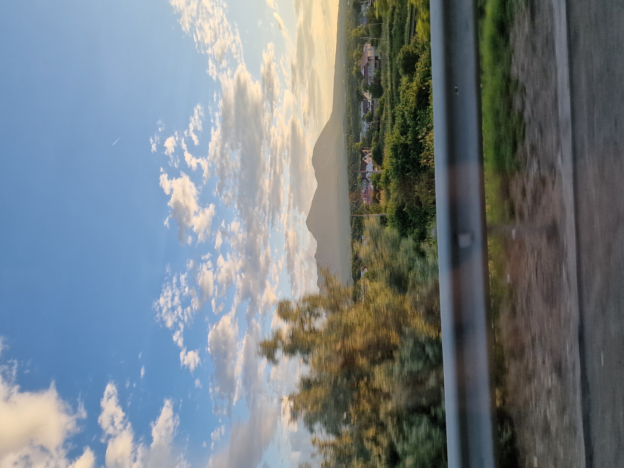 The image depicts a landscape scene with a road in the foreground, separated by a metal guardrail. Beyond the guardrail, a village is nestled among green trees and rolling hills, leading to two distinct mountains in the background. The sky above is a clear blue, dotted with scattered white clouds, and a faint contrail from an aircraft is visible. The lighting suggests it is late afternoon or early evening, with a warm glow illuminating the scene. The village appears to be located in the vicinity of Mount Zmeyka, in Zmeika, Russia, given the characteristic double-peaked mountain formation.