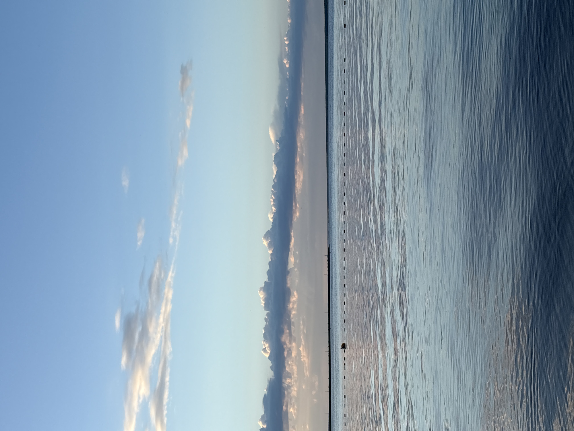This image, correctly oriented as a landscape view, depicts a tranquil waterscape under a vast sky, likely captured in the Chaikovsky, Russia area. The scene is bisected horizontally by a distant horizon.
The upper portion of the image features a wide expanse of clear, light blue sky, with a few scattered, wispy clouds. Along the horizon, a prominent, elongated band of cumulus clouds stretches across the frame. These clouds appear darker and more substantial in their main body but are illuminated with soft, warm, glowing edges, suggesting the presence of either a rising or setting sun just beyond the frame or behind the cloud bank.
The lower half of the image is dominated by a calm body of water, possibly a large river or lake, reflecting the subdued light of the sky. The water's surface is textured with gentle, parallel ripples, creating varying shades of blue, grey, and silver. Near the horizon line, floating on the water, is a small, dark, solitary object, which could be a buoy, a bird, or a small boat. Extending horizontally across the water, precisely along the horizon and parallel to the distant clouds, is a dotted line of small, dark, rectangular shapes. These appear to be markers or buoys, possibly delineating a channel or a boundary on the water.
The overall impression is one of serene natural beauty and peacefulness, characteristic of either dawn or dusk due to the soft, indirect lighting. No people or visible text are present in the image.