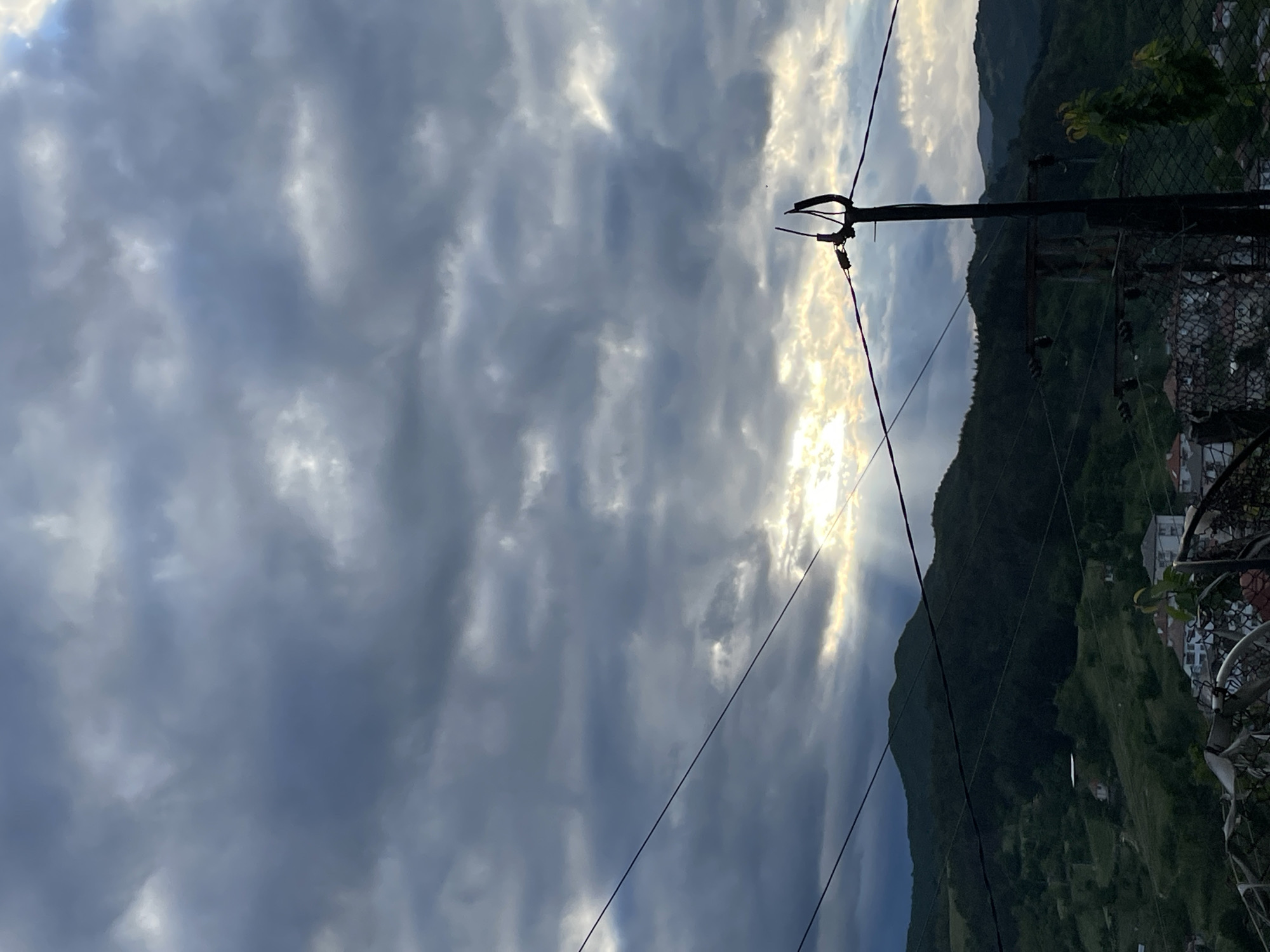 The image captures a view in Zlatograd, Bulgaria, showing a sky dominated by layered, grey clouds partially obscuring the sun. A power line stretches across the frame, running from left to right. The right side of the image reveals a hillside covered in vegetation, and a partially visible section of a residential area. A fence and some foliage are visible in the immediate foreground on the right.