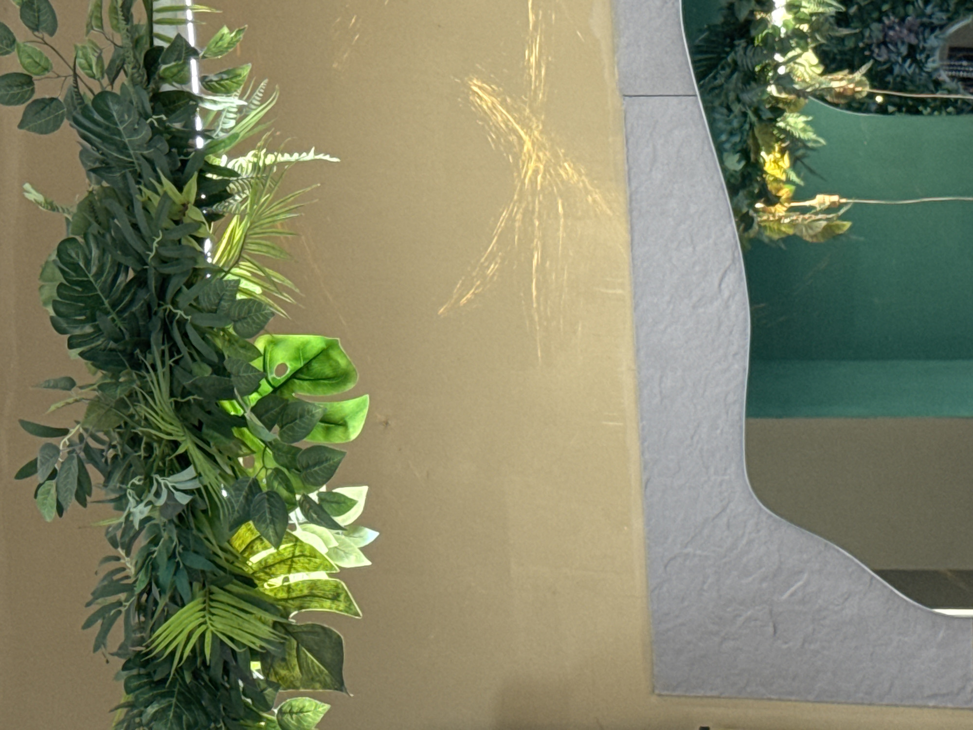An interior scene within a building in Chaikovsky, Russia, showcases a decorative arrangement dominated by artificial foliage. In the upper third of the image, a dense canopy of various imitation green plants, including large-lobed leaves, ferns, and ovate leaves, hangs overhead, illuminated from behind by a hidden light source that casts bright green light through translucent leaves. The middle section features a plain, light beige-colored wall, upon which narrow, golden, wavy light reflections are prominent, forming a distinct 'V' or 'X' pattern. The lower third reveals the top portion of a mirror framed by an irregularly shaped border with a light grey, textured surface. The mirror's reflection shows a continuation of the artificial greenery against a dark teal background, with thin white wires visible, likely part of the lighting or suspension system for the foliage. No individuals are present in the frame.