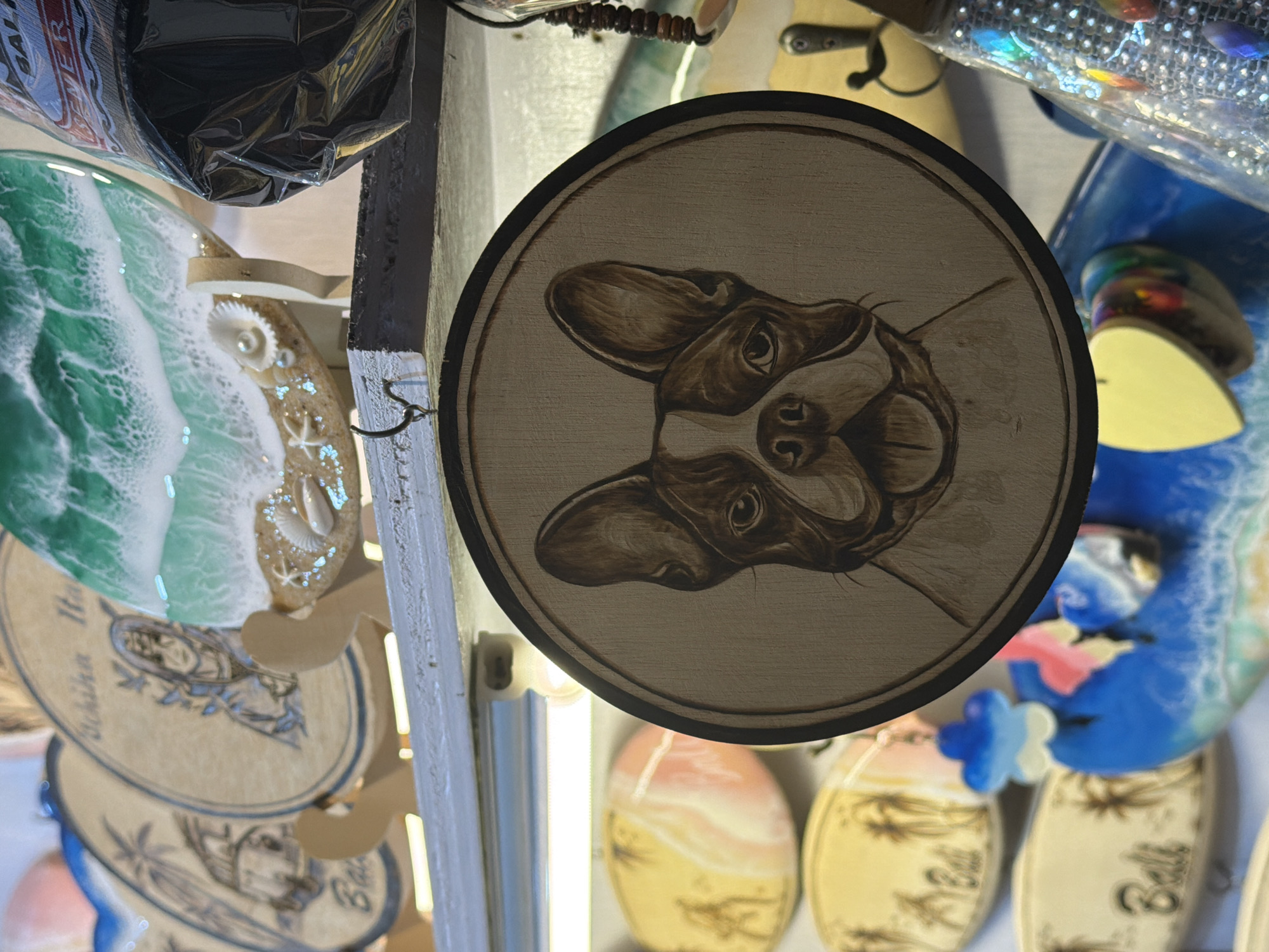 This media file displays a collection of decorative items, likely souvenirs or art pieces, in a market or shop setting in Kuta, Indonesia. The central subject is a round wooden plaque featuring a pyrography (wood-burning) portrait of a French Bulldog's face. The dog has large, expressive ears and dark, detailed markings.
Surrounding the dog plaque are other decorative items. To the left, a round resin art piece depicts a stylized ocean wave with shades of green and white, and embedded seashells and starfish on a sandy background. Below this, several oval wooden pieces are visible, some featuring etched designs of palm trees and what appear to be stylized figures. One of these oval pieces has visible text, though it is partially obscured and difficult to read clearly, but fragments like "Achika" and "Ball" are discernible.
In the background, more colorful, abstract resin art pieces can be seen, predominantly in blue and yellow tones, some with playful, cloud-like embellishments. A string of decorative beads with iridescent elements is also visible at the top right. The lighting suggests an indoor setting, possibly with artificial lights illuminating the products. The overall impression is of a vibrant display of artisanal crafts, catering to a tourist market with themes of beach life and pets.