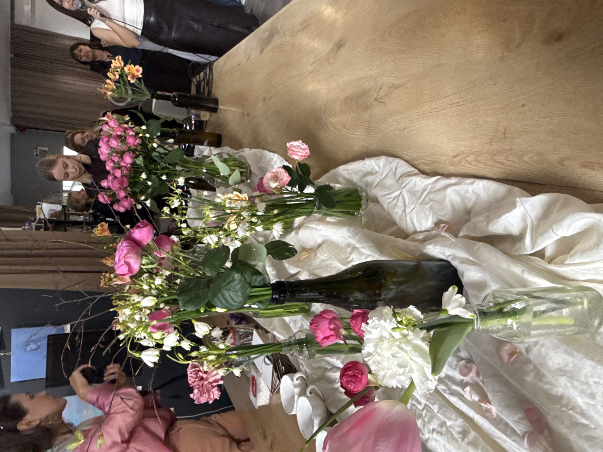 The image captures an indoor scene with floral arrangements dominating the foreground, set against the backdrop of people and furniture.
In the foreground, various types of flowers, including roses, tulips, carnations, and small white flowers, are arranged in different containers, some of which appear to be repurposed wine bottles and glass vases. A rumpled white cloth forms the base for the floral arrangements.
In the background, several people are visible. One woman stands, holding a microphone. Other people are seated or standing near a wooden table. The overall setting suggests an informal gathering or event.
The location, Batumi, Georgia, is not visibly evident from the image itself, though this information has been provided. There are no discernible landmarks or features that would explicitly identify the location.