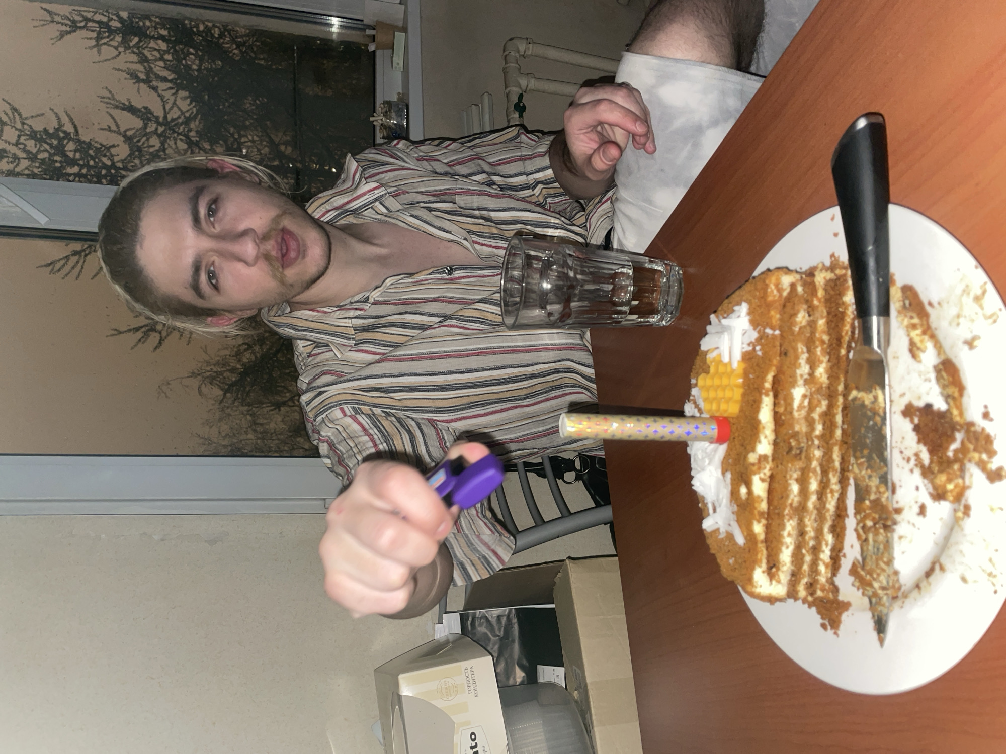 The image captures a young man indoors, seen from a high angle, in the process of lighting a celebratory item on a cake. The man, with light-colored hair pulled back and a mustache, wears a striped button-down shirt and looks directly at the camera with a slightly pursed expression. He holds a purple lighter in his right hand, aiming it towards a small, patterned party popper or candle stuck into a partially eaten cake.
The cake, resting on a white plate on a wooden table, is visibly layered with brown cake and white cream, resembling a honey cake (Medovik). A knife with a black handle is plunged into the cake, suggesting slices have already been taken. A glass containing some liquid is also on the table next to the man's left hand.
In the background, a window reveals the silhouette of tree branches against a muted, light brown sky, indicating either dusk or night-time. Industrial-style pipes are visible along the wall behind the man. Below the table, there are cardboard boxes, one of which has Cyrillic text that reads "Гордость кондитера" (Gordost konditera), meaning "Confectioner's Pride," confirming the Moscow, Russia location context. The scene suggests an intimate celebration, possibly a birthday, taking place in a casual, home setting during the evening.