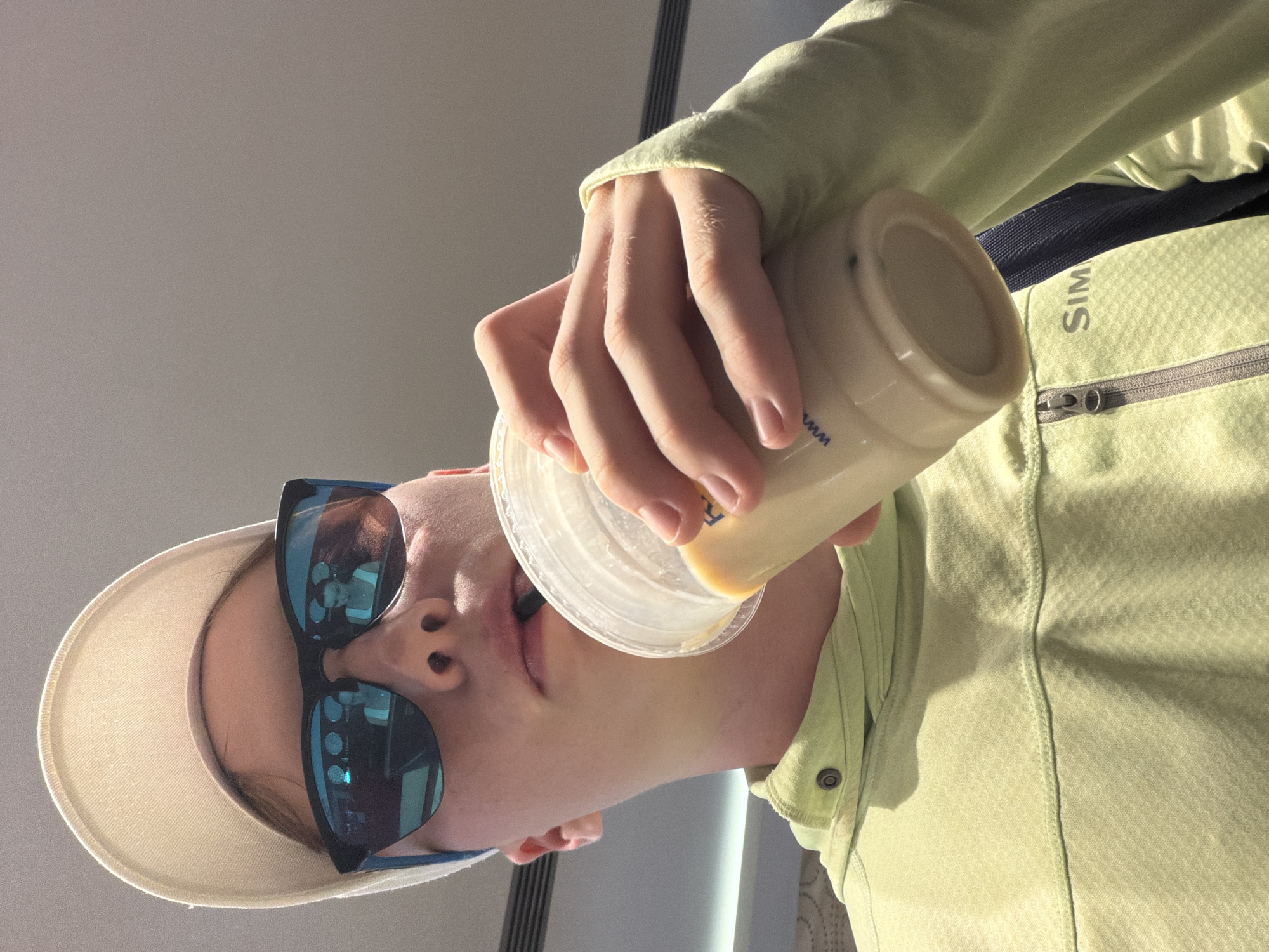 A young person, wearing a light-colored baseball cap and reflective sunglasses, is captured from a low angle while drinking from a large, clear plastic cup filled with a light-colored beverage and ice. They are wearing a lime-green hooded jacket with a zipper and a visible logo that appears to read "SIM". The background is a neutral, indoor setting with a smooth, light-colored ceiling. Reflections in the sunglasses show two other individuals, one with dark hair and wearing a white top, and another with lighter hair also wearing white. The overall scene suggests a casual, possibly urban, environment in Kemer, Turkey.