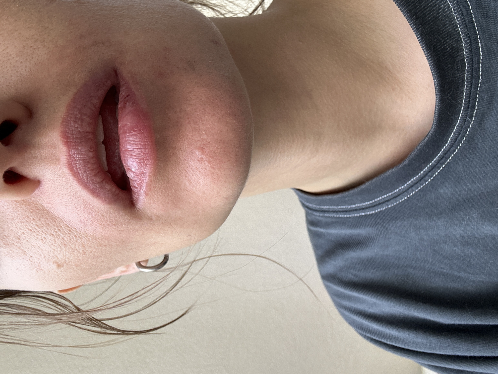 A close-up, waist-up portrait of a person with their mouth slightly open, revealing teeth. The person is wearing a dark gray t-shirt with white stitching around the neckline and sleeves. Their skin appears to have visible pores and some minor blemishes. A small silver hoop earring is visible in their left ear. The background is a plain, light-colored wall. There is no indication of the city of Murino, Russia in the image.