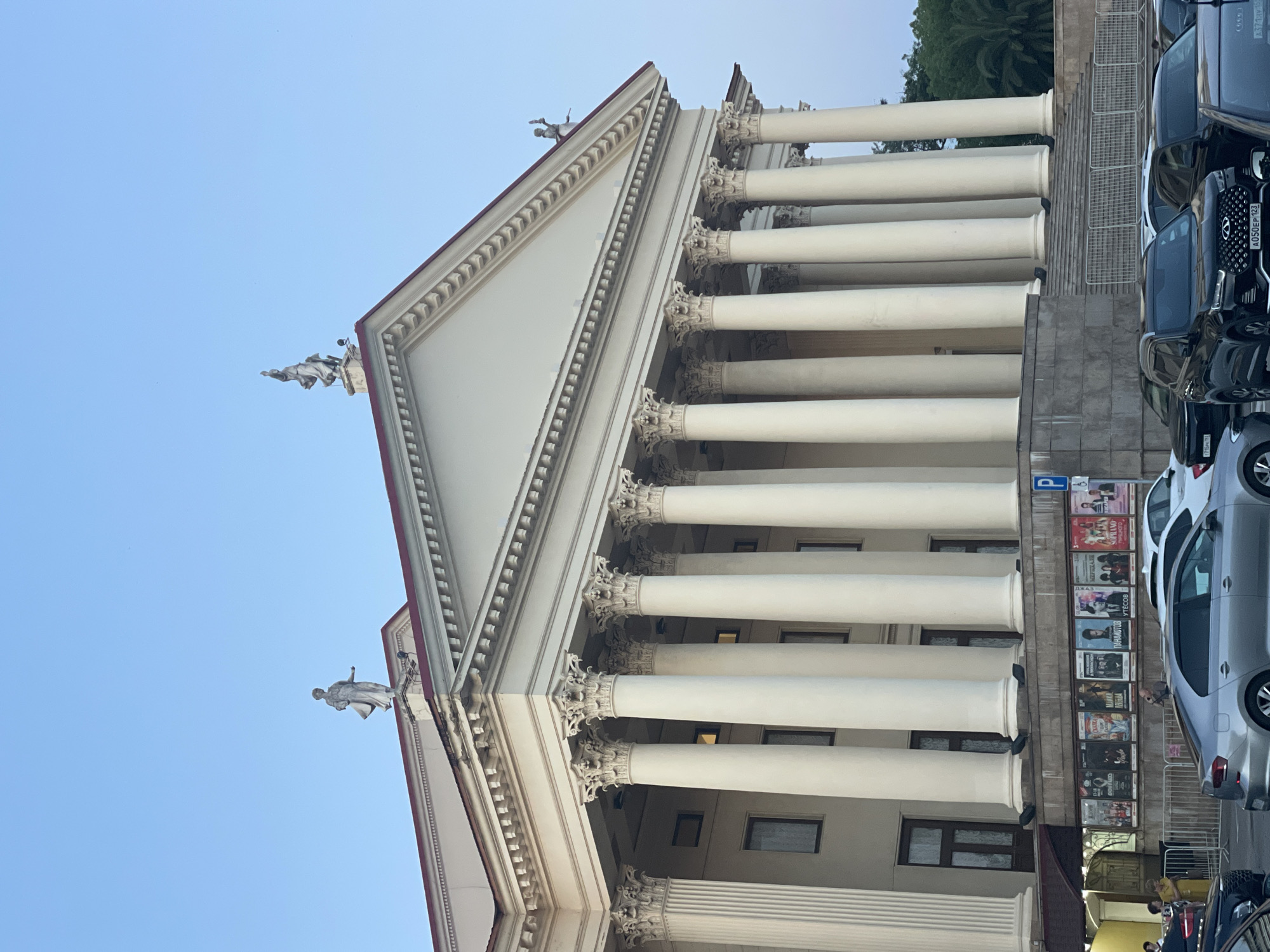 This image depicts the Sochi State Circus, a prominent landmark in Sochi, Russia. The photograph captures the building's grand Neoclassical facade, characterized by a series of tall, white Corinthian columns supporting a pediment. Sculptures of classical figures adorn the roofline. In front of the circus, posters advertising various performances are displayed on a facade. A parking lot filled with cars is visible in the foreground, suggesting a public event or arrival of visitors. People can be seen near the entrance of the building, some walking and others standing. The sky is clear and blue, indicating it's daytime. The overall scene conveys a sense of a vibrant public space.