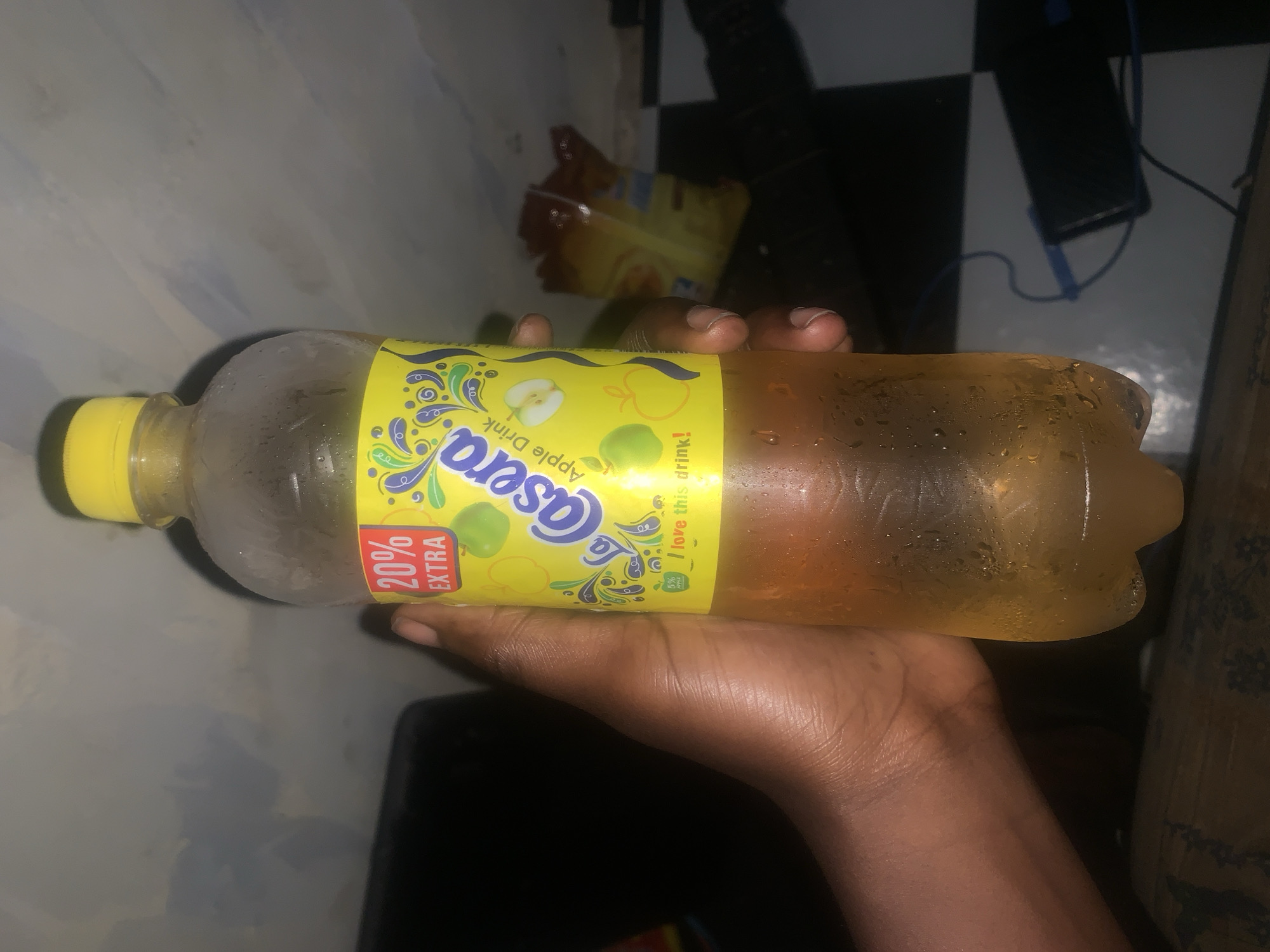 A photograph depicts a hand holding a bottle of Casera Apple Drink. The bottle, visibly cold with condensation, is slightly tilted, displaying its yellow label with the Casera logo and apple illustrations. A partial view of a snack bag is also visible, and the background appears to be a residential setting in Rafin Par, Nigeria, indicated by the combination of objects and cultural context.