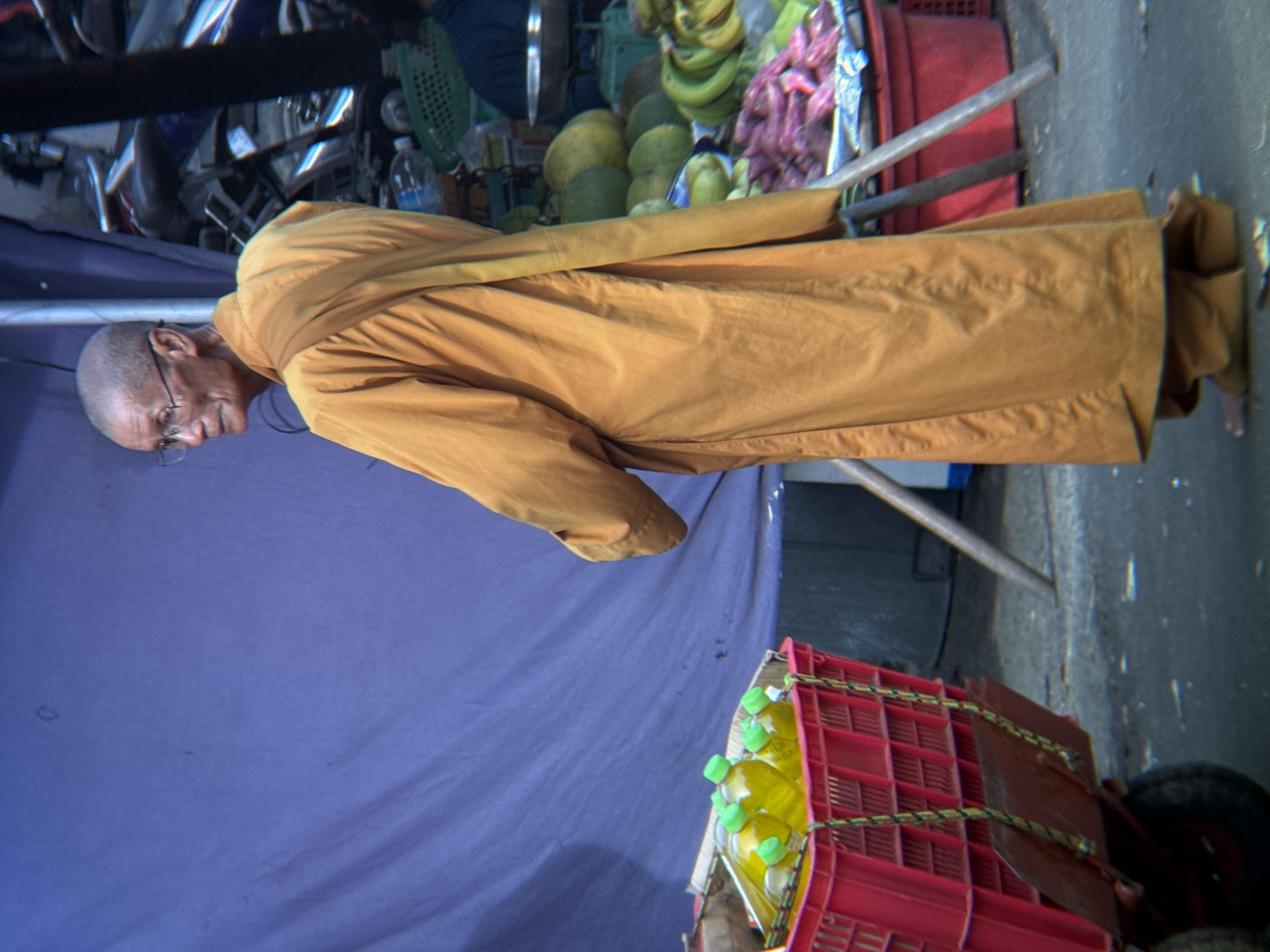 A Buddhist monk wearing a saffron robe stands in a market setting in Nha Trang, Vietnam. The monk is bald, wears glasses, and is looking away from the camera. In the foreground, a red plastic crate on a cart holds several bottles of yellow liquid with green caps. Behind the monk, various fruits and vegetables are displayed, including what appear to be bananas and sweet potatoes, in red and green baskets. A large purple tarp is visible in the background, likely part of a market stall. The ground is paved with a rough texture, and the overall lighting suggests it is daytime.