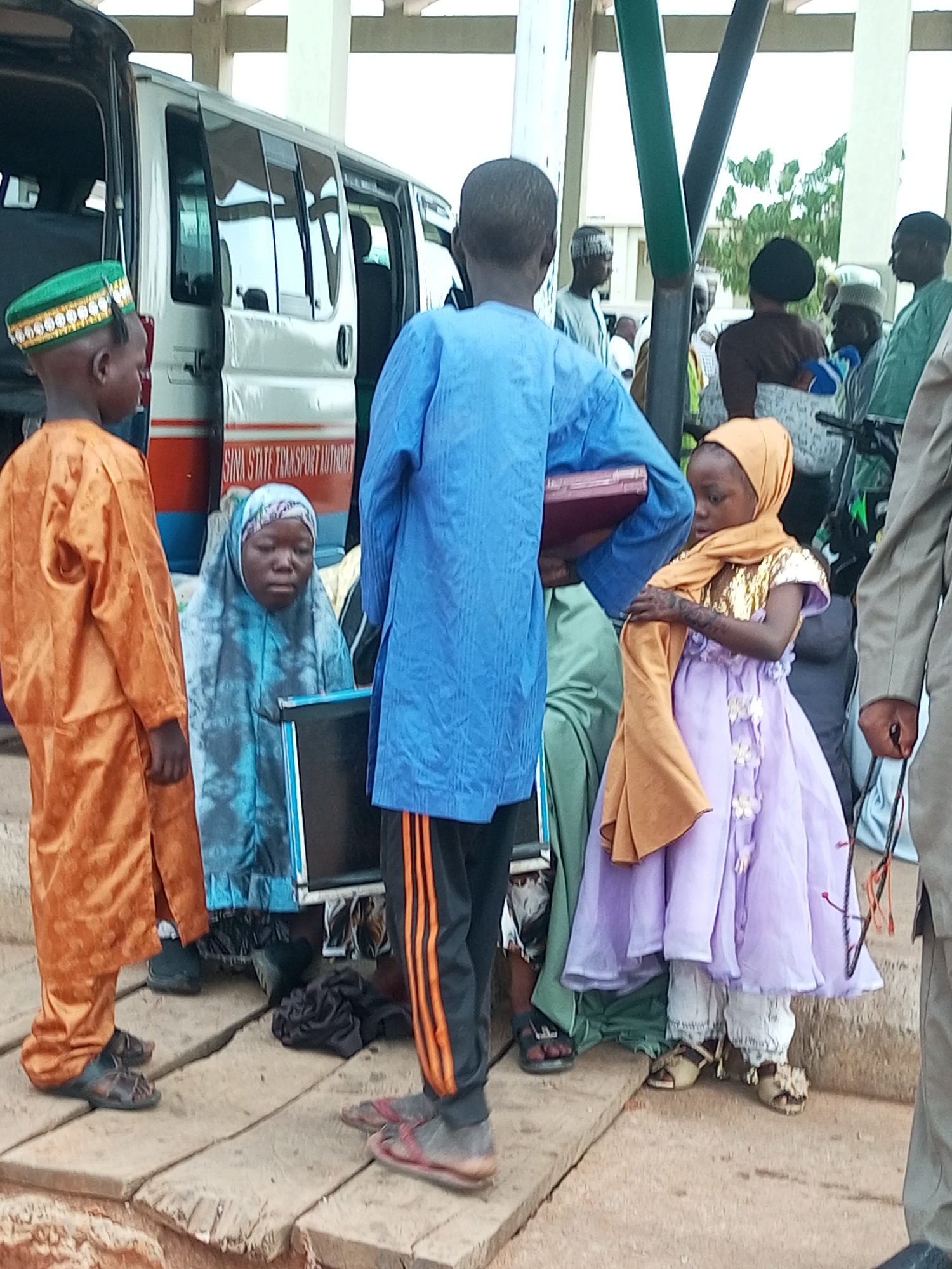 The image shows a group of people, including children, gathered near a bus in what appears to be a transportation hub in Gombe, Nigeria. The bus, labeled "GOMBE STATE TRANSPORT AUTHORITY," has its doors open, suggesting people are boarding or disembarking.
In the foreground, a young boy in an orange robe and green cap stands to the left, facing away from the camera. Next to him, a girl in a blue hijab sits, looking downwards. A young girl in a purple dress and orange hijab stands to the right, holding a shawl. Several other individuals, mostly men in traditional attire, are visible in the background, some with their backs to the camera.
The ground is made of rough wooden planks, and the environment suggests an outdoor setting, possibly a bus station. The lighting indicates it is daytime. The overall scene depicts a moment of activity and waiting within a community context.