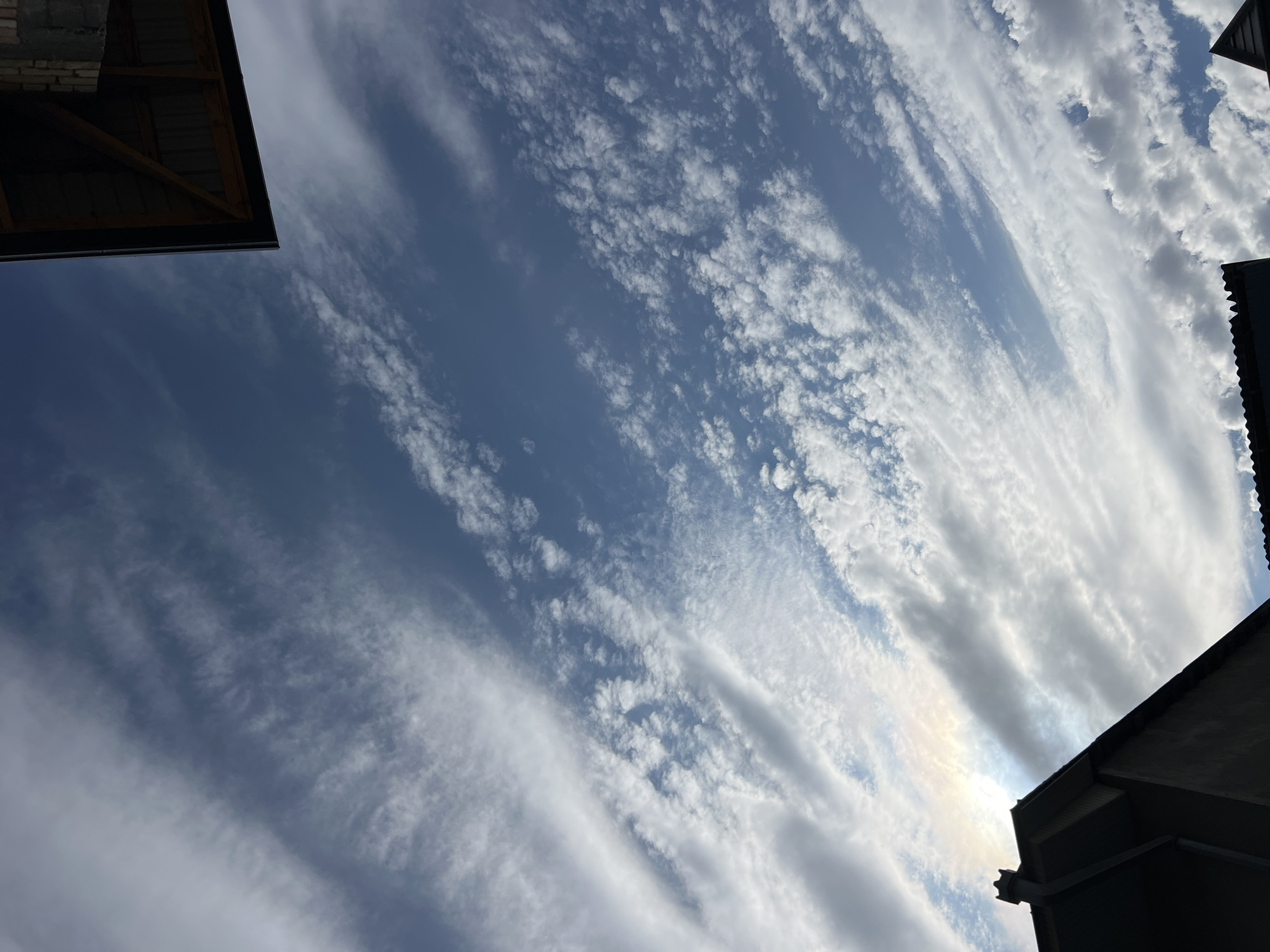 The image is a low-angle, outdoor shot of a cloudy sky. The sky is a mix of blue and white, with wispy clouds stretching across the frame. In the foreground, the dark silhouettes of two rooftops are visible. One rooftop is in the lower left corner, a dark angular shape with a decorative peak. The other rooftop is in the upper right corner, showing the underside of a sloped roof with wooden beams. The sun is partially obscured by clouds, creating a bright, hazy area in the sky near the lower left rooftop. The image appears to be taken in an urban or suburban environment.