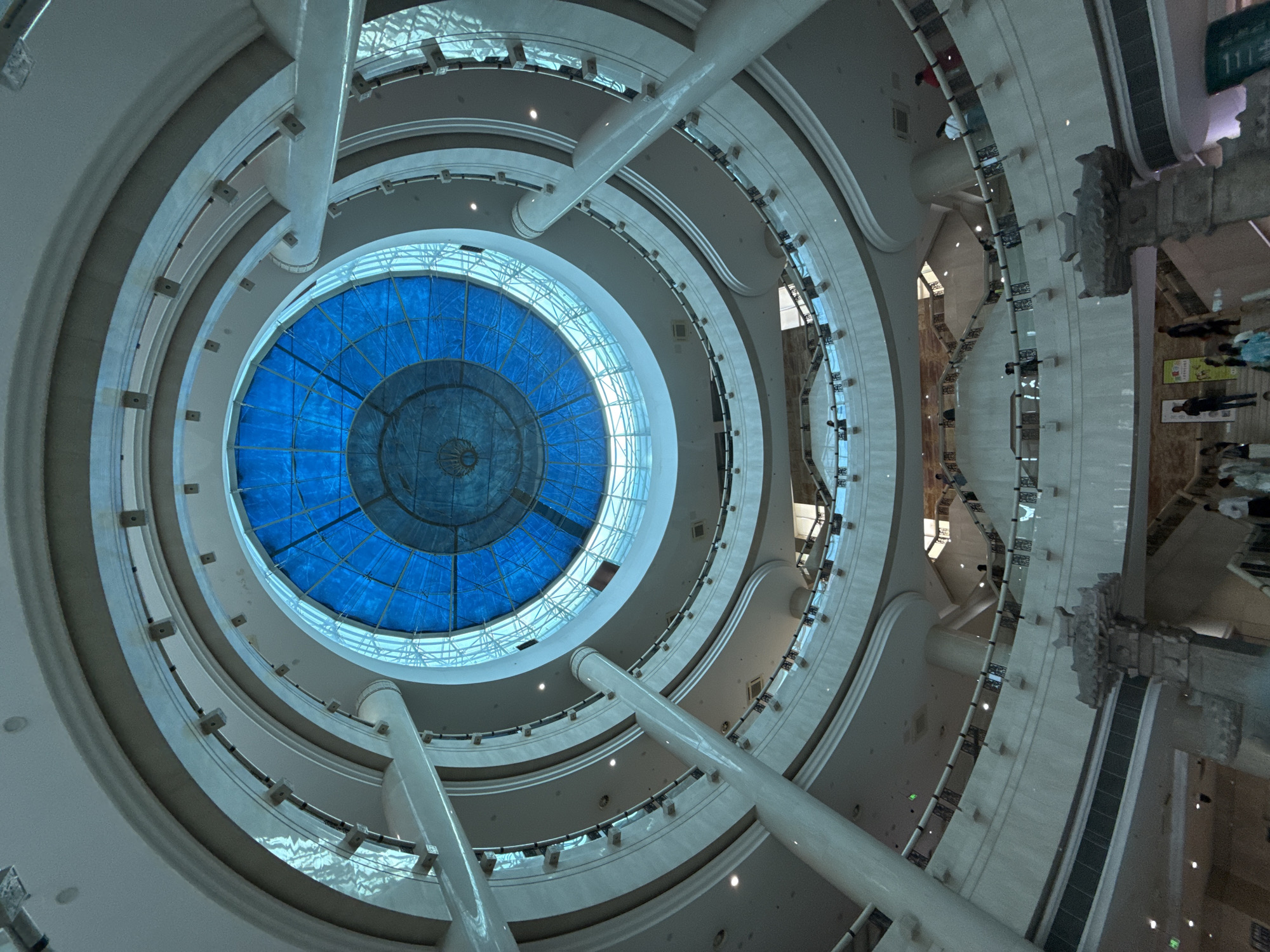A low-angle, interior shot captures a multi-story atrium with a large, circular, blue glass dome ceiling. White, curved balconies with black railings line each level, supported by large, cylindrical white pillars. Several people are visible on the balconies and staircases, appearing to be shopping or observing the architecture. The lower levels of the atrium feature decorative stone pillars and some posters are displayed on the walls. The scene appears to be in a modern shopping mall or public building in Yuzhong District, China.