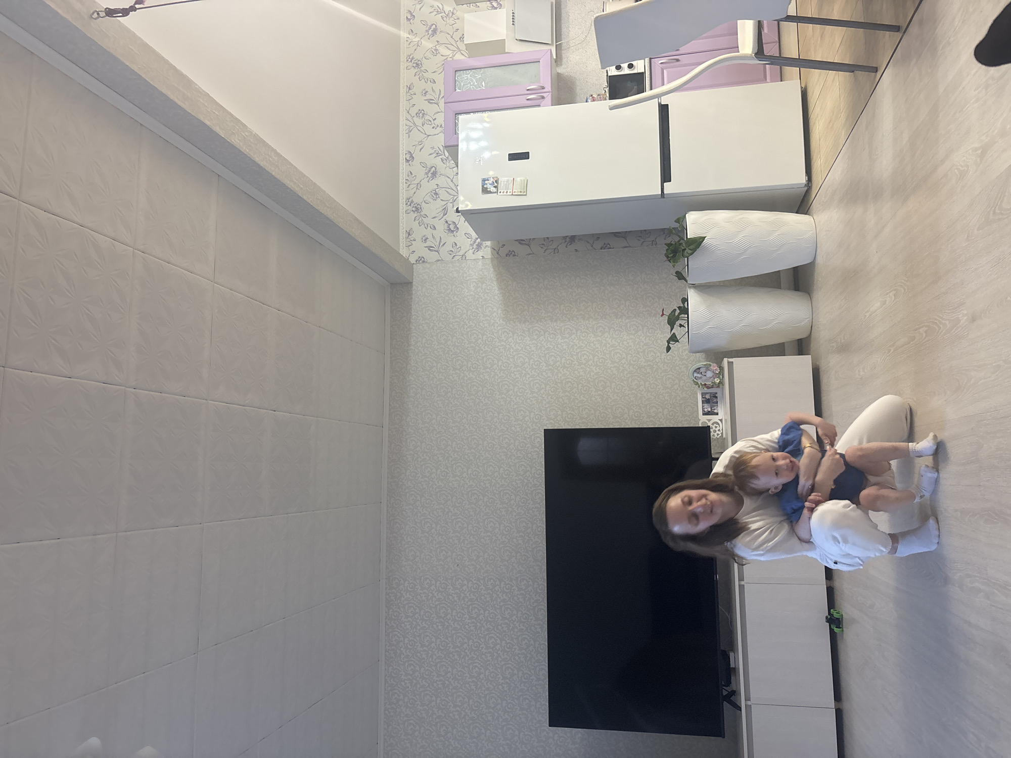A woman holds a toddler in her arms, both seated on a light-colored floor in what appears to be a home interior, possibly in Nestiukovo, Russia. The room includes kitchen appliances such as a refrigerator, as well as a TV and shelving unit. Wallpaper with a patterned design is visible on the walls.