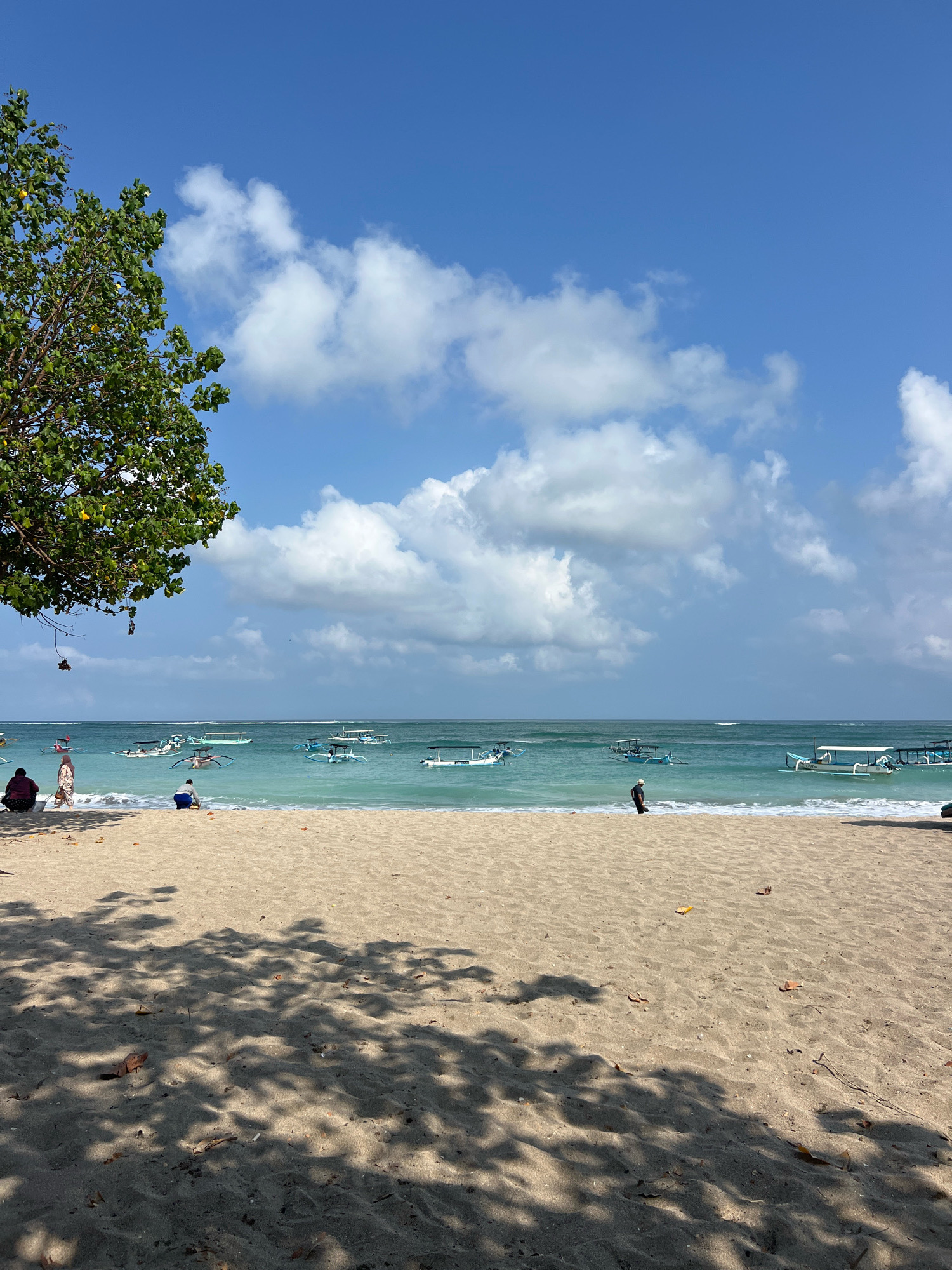 A wide shot captures a tranquil beach scene in Kuta, Indonesia. In the foreground, golden sand is dappled with the shadows of unseen foliage, with a few scattered leaves and twigs. The midground features the turquoise ocean, dotted with numerous traditional Balinese jukung boats, some of which are stationary while others appear to be moving gently. Along the shoreline, three individuals are visible. On the left, two women are seated on the sand, one wearing a headscarf. Further to the right, a person is crouched down near the water's edge. A lone bird stands on the beach to the right of the frame. In the background, the vast expanse of the sky is a brilliant blue, adorned with fluffy white cumulus clouds. The overall impression is one of peaceful seclusion and natural beauty.