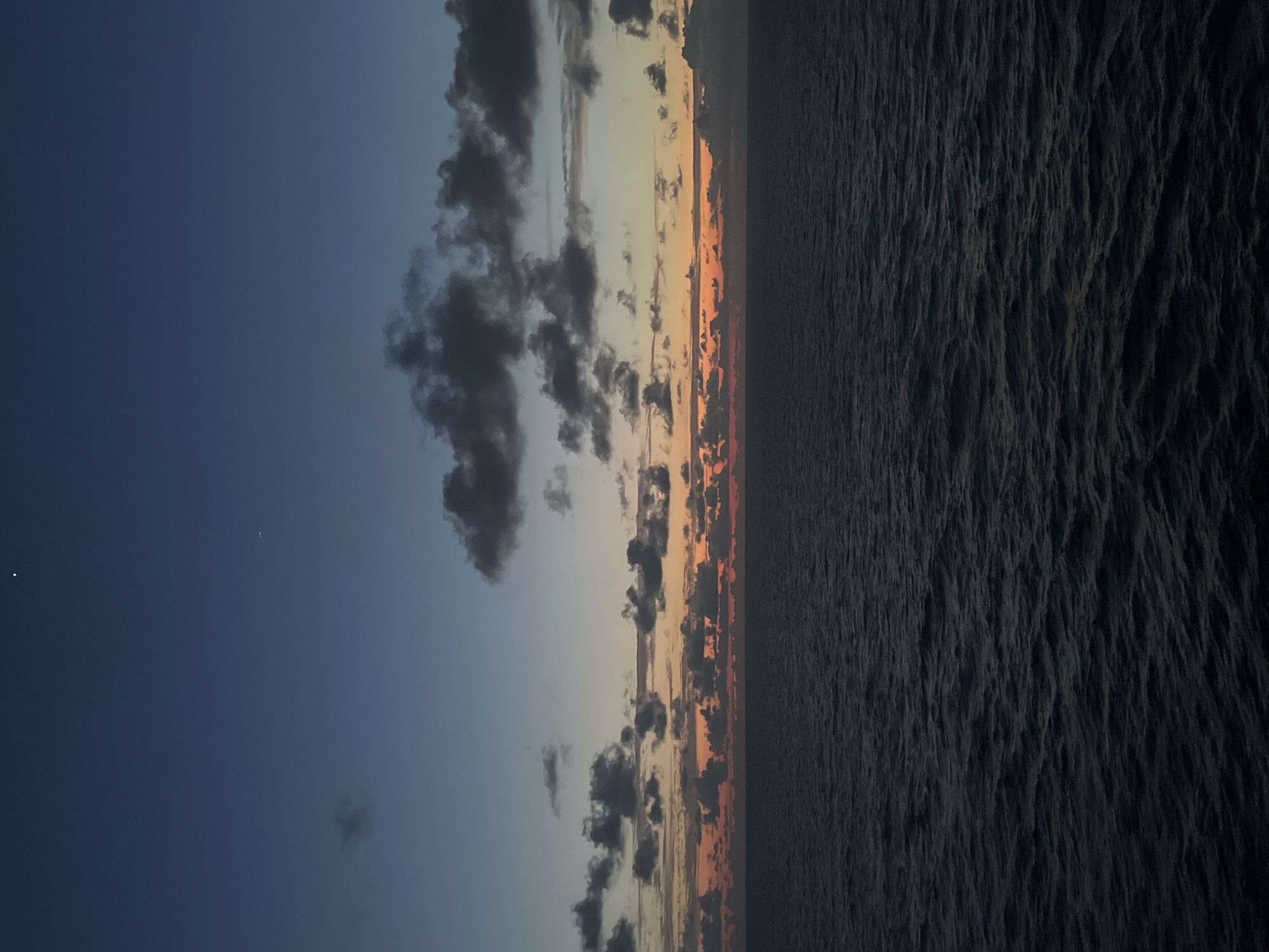 The image depicts a seascape at dusk, with the horizon dividing the dark, rippling ocean from a twilight sky. Scattered clouds are present in the sky, some appearing dark and dense, while others are lighter and more diffuse. A band of orange and pink hues is visible along the horizon, indicating the setting or rising sun. Two small, bright points of light, likely stars or planets, are visible in the upper portion of the dark blue sky. The overall impression is one of a vast, open body of water under a dim, colorful sky. The location is described as Unknown, Unknown, suggesting it is not within a specific city or known geographical area.