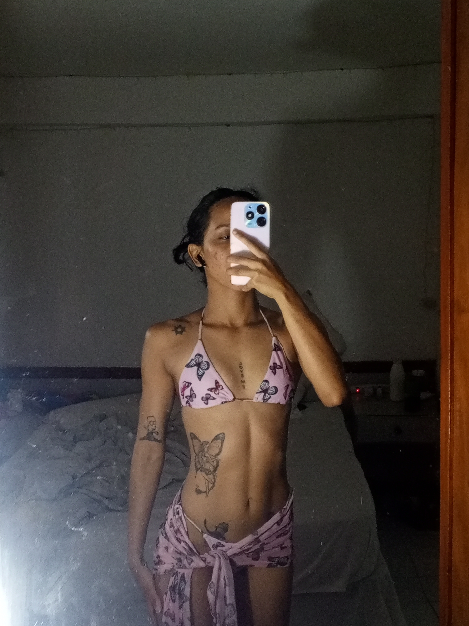 A person is standing in front of a mirror, taking a selfie. The person is wearing a pink bikini top and sarong with butterfly prints. The person is holding a phone in front of their face, obscuring the face. There are tattoos on the person's arm, stomach, and collarbone. The room is lit by natural light and the location could be Porlamar, Venezuela. A bed with rumpled sheets is visible in the background. A bedside table with bottles on it can also be seen.