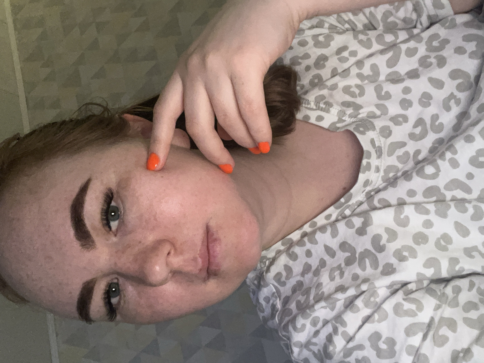 A woman with auburn hair and fair skin is lying down, facing the camera at an angle. She is wearing a white t-shirt with a grey animal print. Her right hand is raised, with her fingers touching her face near her cheek. She has green eyes and freckles. Her fingernails are painted bright orange. The background is a light grey surface with a subtle geometric pattern. The location in the city of Chaikovsky, Russia, is not discernible from the image.