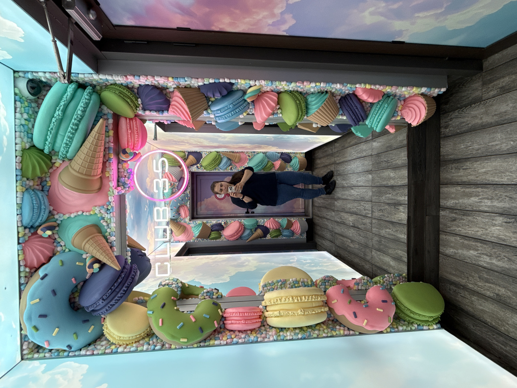 The image depicts an interior space with whimsical dessert-themed decorations. The walls are adorned with oversized, colorful replicas of macarons, donuts, ice cream cones, and other confections, embedded among small, pastel-colored spheres resembling sprinkles or candies. A person is standing in the center of the space, holding a smartphone and appearing to take a picture of themself.
The perspective is unique, as if the room is a vertical shaft or tunnel, with the floor and ceiling lined with the dessert decorations. Above and below the decorative elements are projected images of sky and clouds. The word "CLUB" is visible in neon lights on the left wall.
The location is Sofia, Bulgaria.
