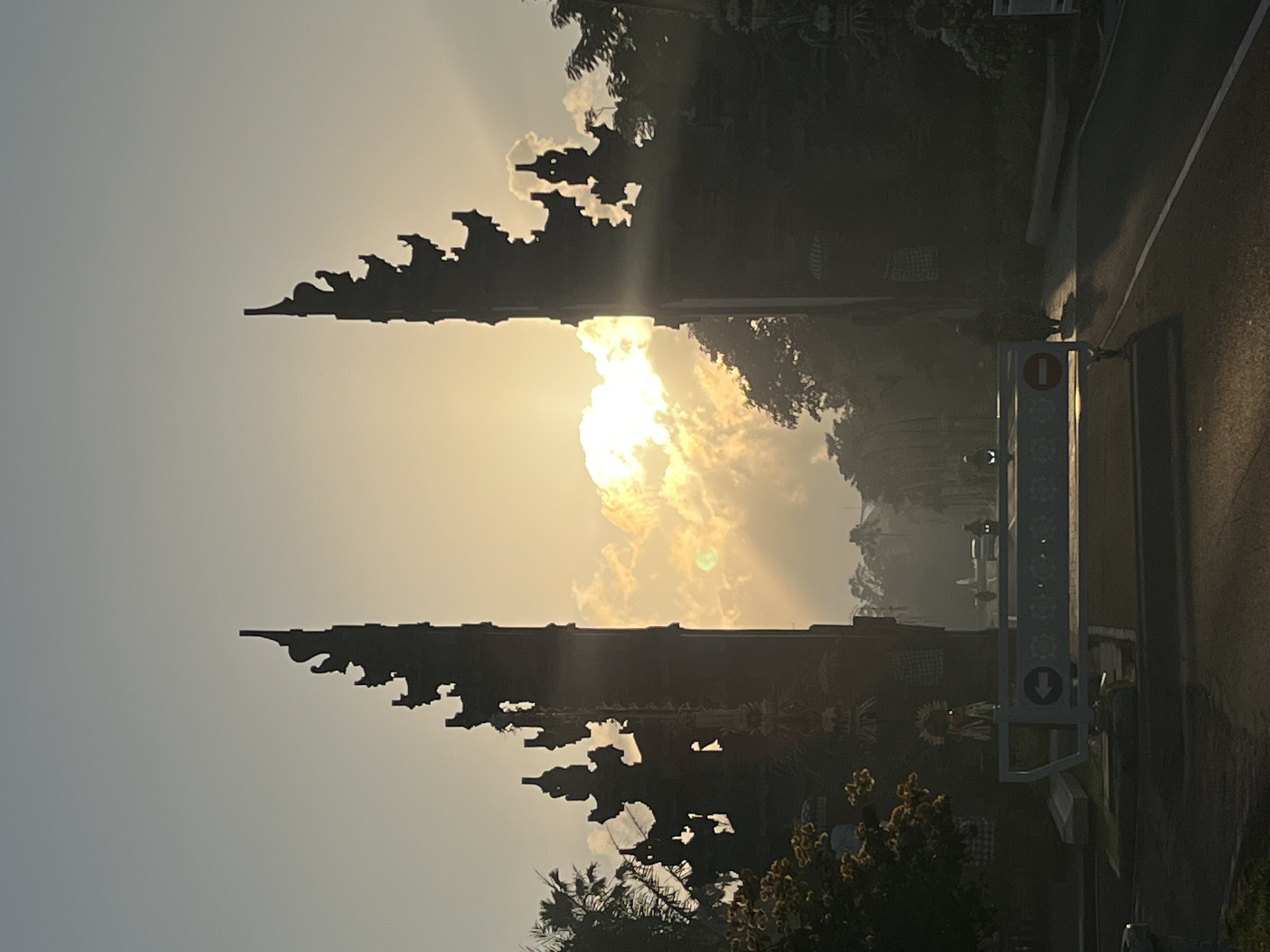 The image captures the silhouette of a Balinese split gate, known as Candi Bentar, at sunrise in Kuta Selatan, Indonesia. The sun is positioned behind the gate, casting dramatic rays through the clouds, illuminating the scene with a warm, golden light. Two towering, ornate gate structures frame the view, their intricate details softened by the backlight. In the foreground, a road is visible, with a temporary barrier or gate. A white barrier with a blue arrow and text indicating direction is present. A person on a motorcycle is visible in the distance, approaching the gate. Trees, including palm trees, line the sides of the road and the area surrounding the gate. The sky is a gradient of pale blue to yellow, indicating the early morning hours.