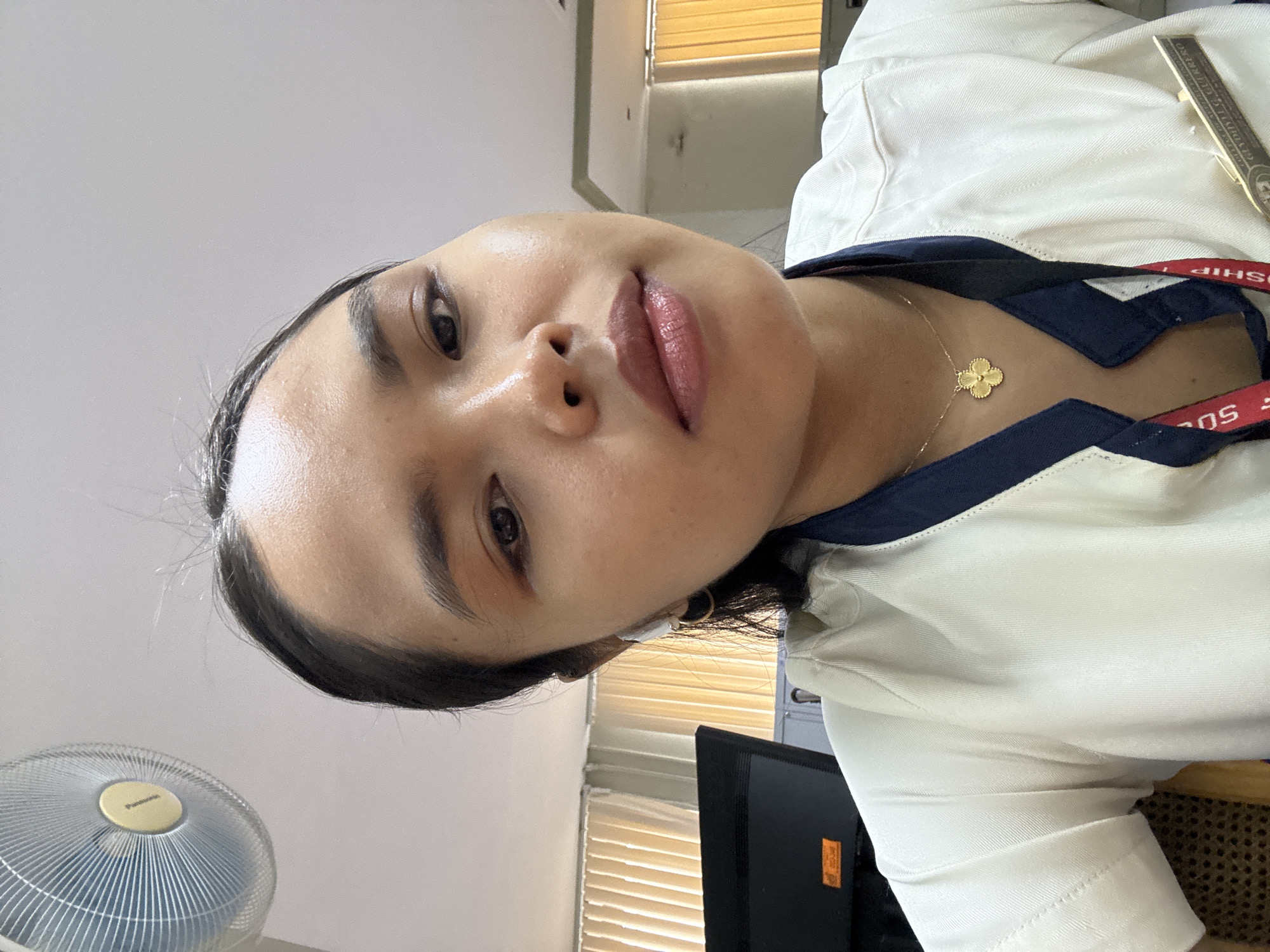 The image is a close-up selfie of a young woman with dark hair, wearing a white collared shirt with a navy blue trim. She has a gold clover-shaped pendant necklace and is wearing small hoop earrings and a white wireless earbud. A red lanyard with text is visible on her shirt, along with a name tag. The background includes a desk with a monitor and a fan mounted on the wall. The lighting suggests an indoor environment, possibly an office or classroom. The scene appears to be in Davao City, Philippines, as indicated by the name tag on her uniform which reads "GLYDOCELL L. GUERRERO".
