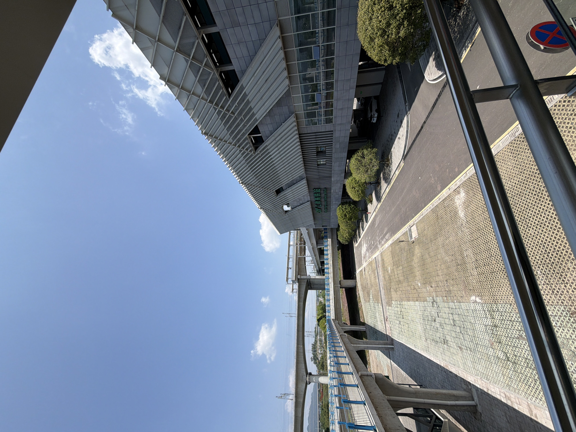The image captures a modern elevated transportation hub and surrounding infrastructure under a bright, clear blue sky with scattered white clouds. In the foreground, a pedestrian walkway with a blue railing curves into the scene, supported by concrete pillars. Beyond this walkway, a multi-level elevated roadway or bridge system is visible, with concrete structures and overhead power lines stretching into the distance.
To the right, a contemporary building with a distinctive angled, grated roofline and a facade of gray tiles and large windows dominates the mid-ground. Green trees line the front of this building, and a street with parked cars is visible beneath an overhang. The ground in the open area between the walkway and the building is paved with interlocking concrete blocks, showing a transition in color indicating different paving materials or wear.
The overall impression is one of urban development and efficient transit infrastructure. The specific location, identified as Jiangjin District, Chongqing, China, suggests this is likely a train station or a major bus terminal designed for high-capacity passenger movement. The clean lines, modern materials, and bright lighting point to a well-maintained and functional public space. There are no people visible in the image, focusing the scene entirely on the architectural and infrastructural elements.