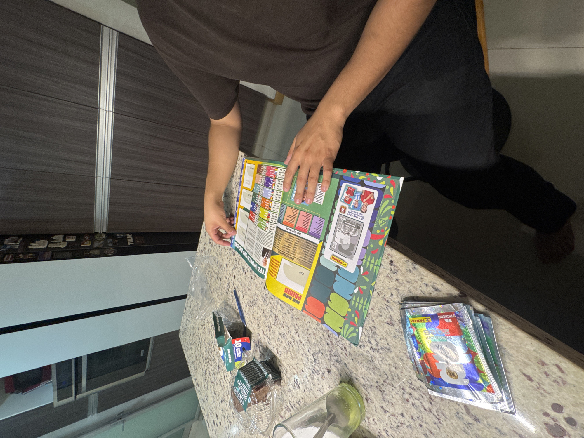 This indoor image, captured in what appears to be a kitchen or dining area in Puebla, Mexico, shows a person engaging with a Panini sticker album. The person, dressed in a dark brown t-shirt and dark pants, is seen from the waist up, with their hands holding open a large sticker album on a speckled granite-patterned countertop. One foot is visible near the bottom right.
The central activity involves the person interacting with the "FIFA WORLD CUP 2026" Panini album. Several unopened Panini sticker packs, prominently displaying "7 STICKERS" and "PANINI" along with "HOANG CUP," are stacked on the right side of the counter. Next to them, there's an empty plastic wrapper, a blue pen, and a small blue box marked "10," suggesting other items have been opened. A glass jar or cup with a spoon inside is also on the counter.
Notable details include the visible text on the album: "FIFA WORLD CUP 2026," "WE ARE PANINI," "MYPANINI," "The Symbols," and "Roll of Honour." Partially visible text includes "GET THE APP" and "YOUR TUUM." The background features dark grey, horizontally-striped cabinetry with a vertical silver strip, and a light-colored floor. The scene suggests a casual hobby activity, likely in the daytime given the general brightness, though direct light sources are not visible.
