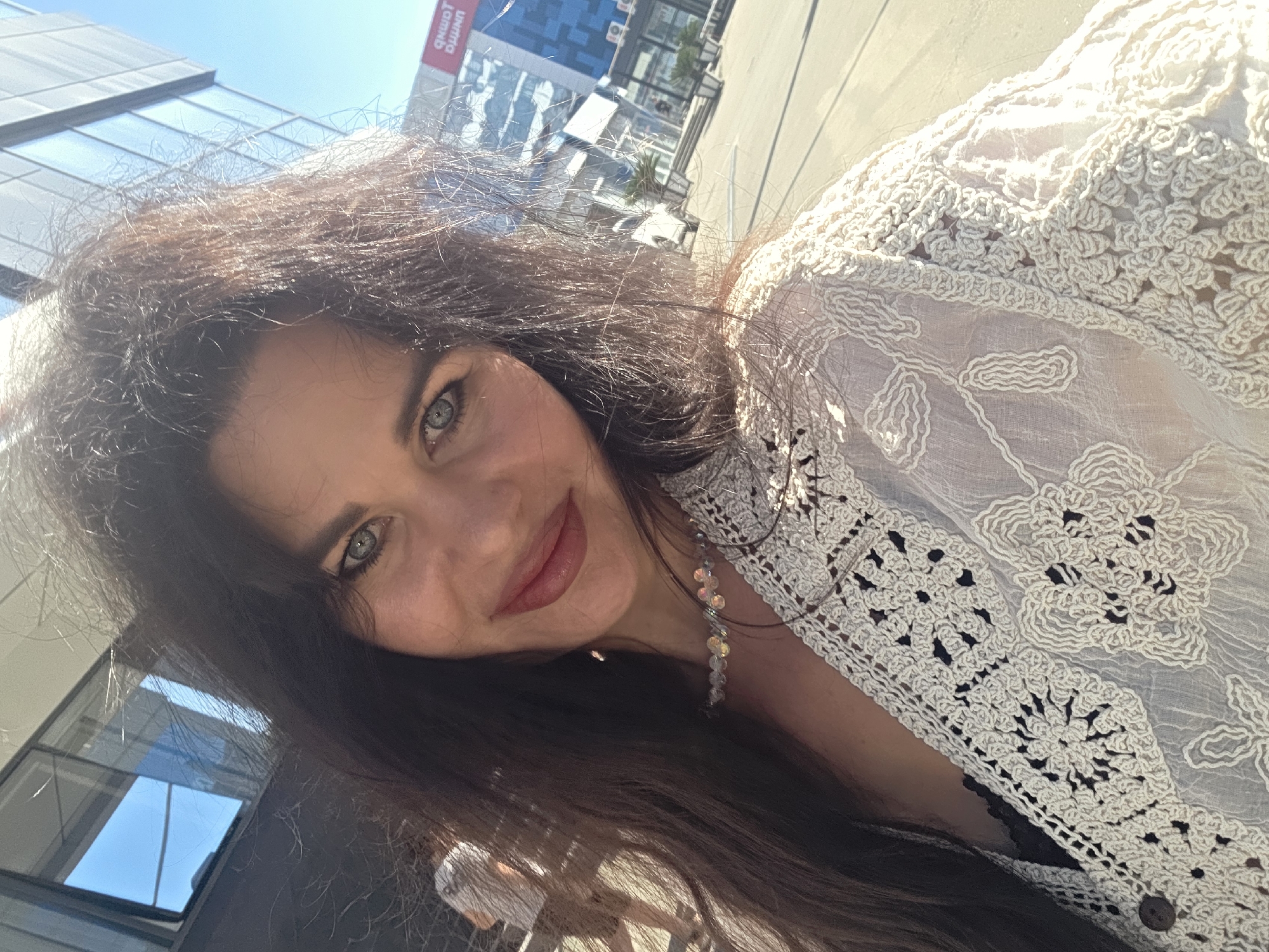 A woman is taking a selfie outdoors. She has long, dark, wavy hair, light blue eyes, and is smiling. She is wearing a white crocheted lace garment over a black top and a beaded necklace. In the background, there is a modern building with glass windows and a blue sky. The scene appears to be a sunny day in an urban environment, possibly a city street or plaza. The presence of a building with reflective glass and what appears to be commercial signage (one sign in Cyrillic script indicating "SOCHI") suggests a location within the city of Sochi, Russia. A blurred figure of another person is visible in the mid-ground to the left.