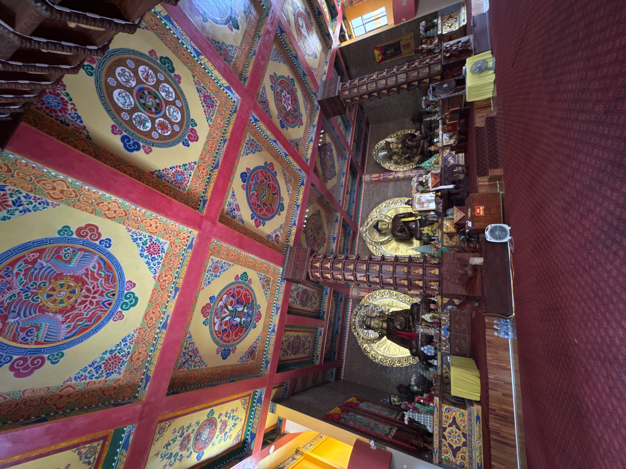 The image shows the interior of a Buddhist temple in Việt Trì, Vietnam. The ceiling is elaborately decorated with colorful paintings, featuring Buddhist motifs such as lotuses, mandalas, and mythical creatures like phoenixes. The walls are lined with small statues and religious artifacts. In the center of the room, three large golden statues of Buddha are prominently displayed on a raised platform. The floor is covered with a deep red carpet. The scene is well-lit, suggesting a daytime view. There are no people visible in the image. The overall impression is one of ornate religious devotion and peaceful contemplation.