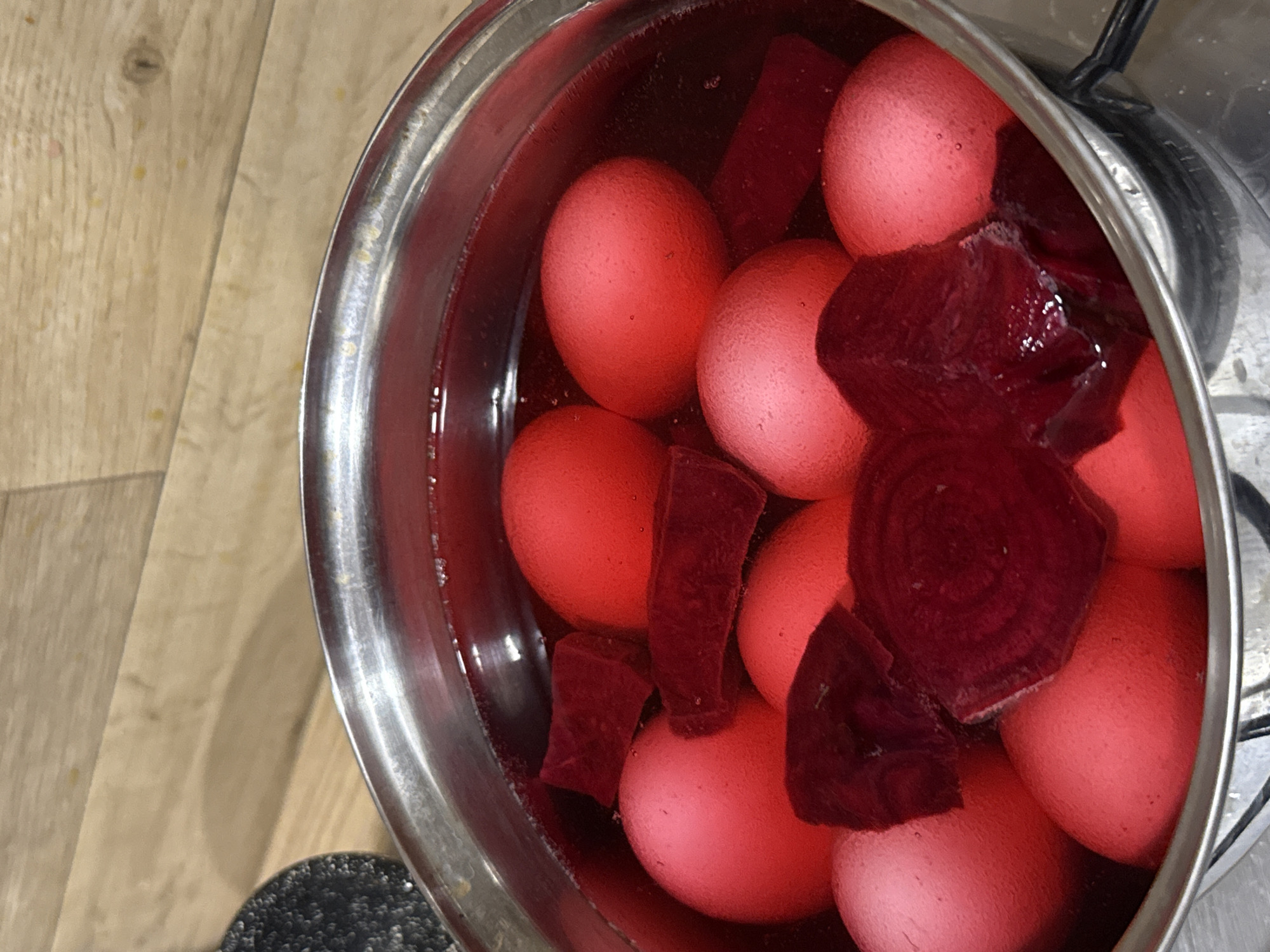 A top-down, close-up shot shows a stainless steel pot filled with pink-tinted hard-boiled eggs and chunks of beetroot. The pot is sitting on a stovetop, likely in a kitchen, although the exact location in Chaikovsky, Russia is not determinable from the image. There are no people visible in the shot. The main subjects are the eggs and beetroot, suggesting a process of dyeing or pickling the eggs. The environment appears clean and domestic, with a wooden countertop partially visible on the left side of the frame.