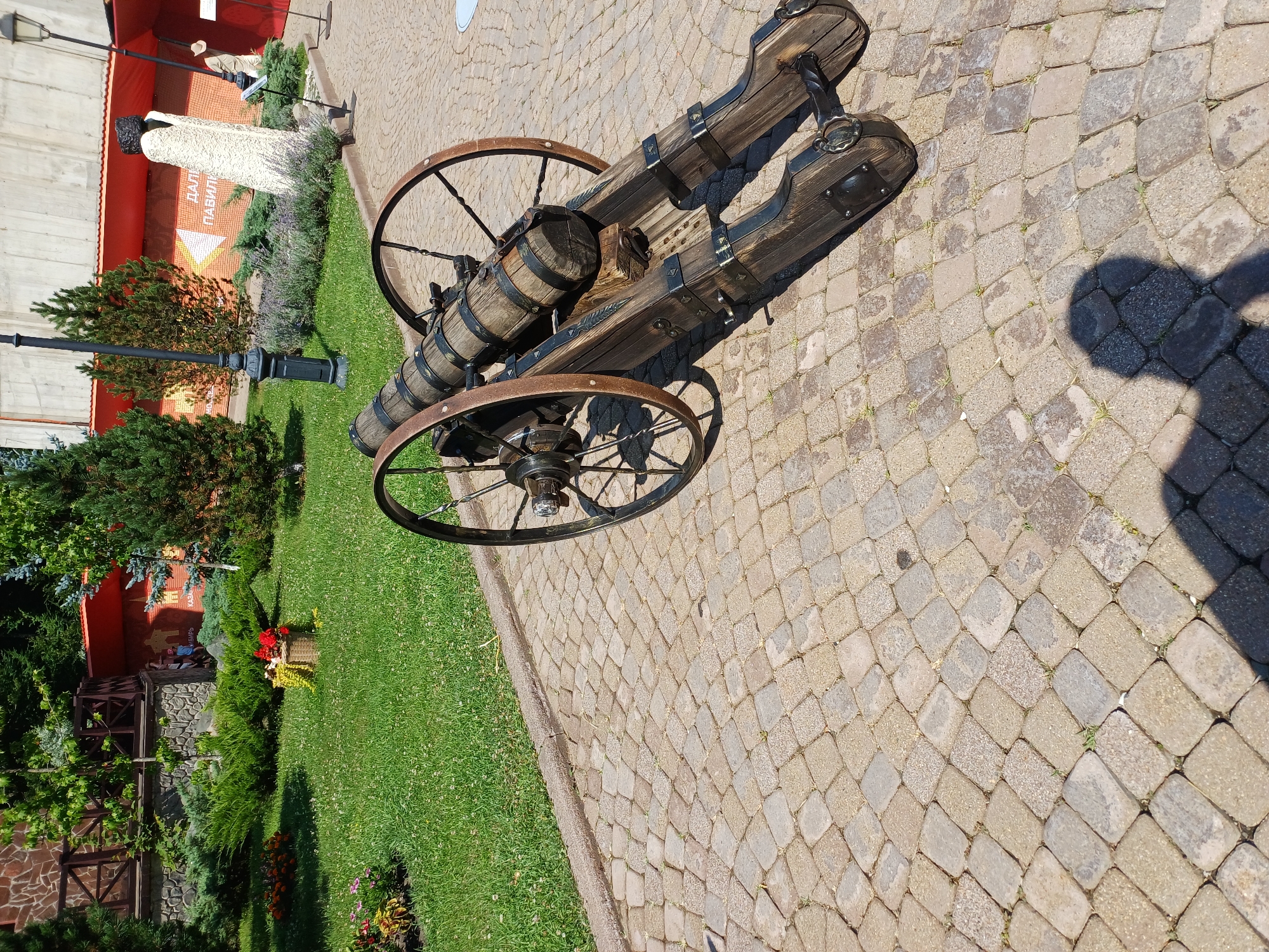 In the city of Estosadok, Russia, a vintage wooden cannon is positioned on a cobblestone path. The cannon, adorned with metal banding, rests on a wooden carriage with two large spoked wheels. Behind the cannon, a well-maintained lawn features a basket of red flowers and various green bushes. In the background, a stone building with a wooden fence and a red awning is visible. Further back, a concrete structure stands next to a statue and a light post. The scene is illuminated by natural daylight, casting shadows on the ground, including that of a person standing in the foreground.