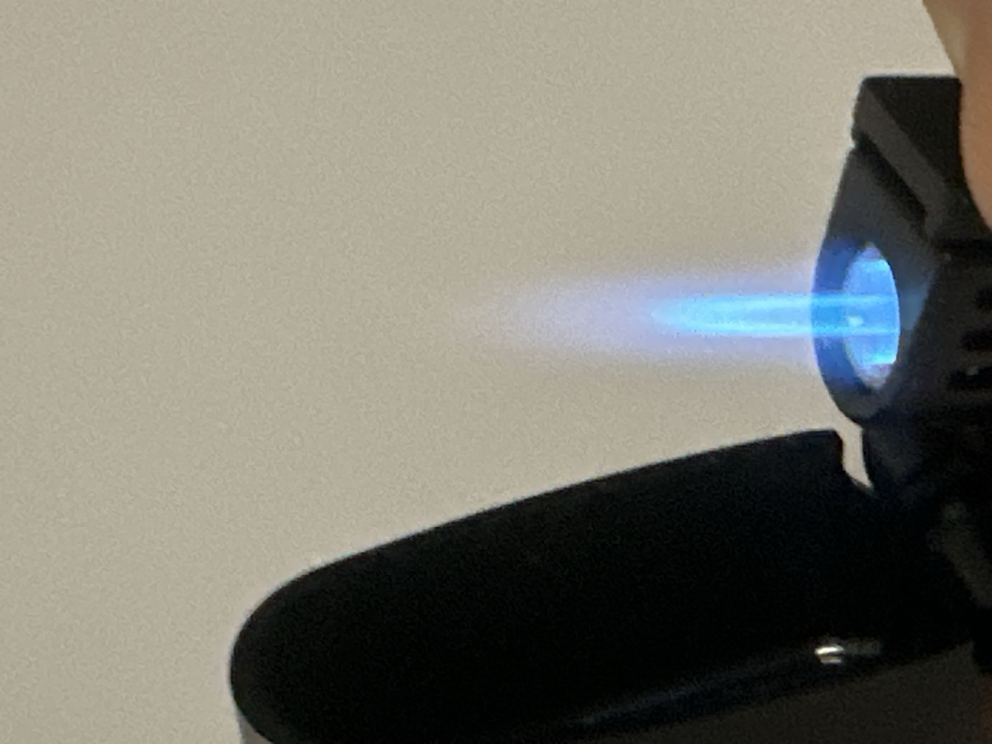 The image shows a close-up of a black lighter with a bright blue flame. The background is a plain, light-colored wall. The lighter is open, and the flame appears to be emanating from a circular opening. The composition is tight, focusing on the lighter and flame, with no indication of any specific location or context beyond the immediate subject. There are no people or other objects visible in the frame. The lighting is somewhat harsh, creating a distinct contrast between the dark lighter and the bright flame. The overall impression is one of a simple, functional object in use, with the flame being the focal point. There is no information in the image to suggest any connection to Sofia, Bulgaria.