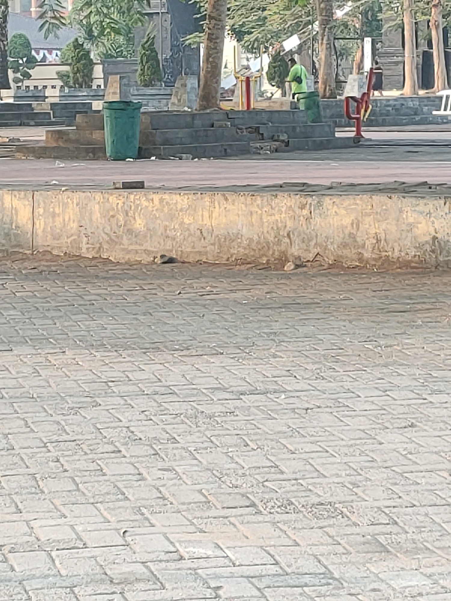 The image depicts an outdoor scene, likely in the city of Kanigoro, Indonesia, focusing on a paved area with a retaining wall and steps leading up to a higher level. In the background, there are trees, buildings, and people. A prominent green trash bin is positioned on the steps. To the right, a person in a bright green shirt appears to be engaged in cleaning activity, pushing a broom towards the trash bin. Further in the background, another person in dark clothing is walking away from the camera. A red metal structure with wheels, possibly a cart or piece of equipment, is also visible near the steps. The ground is covered with light-colored, interlocking paving stones, and the retaining wall is made of concrete with a rough, textured surface.