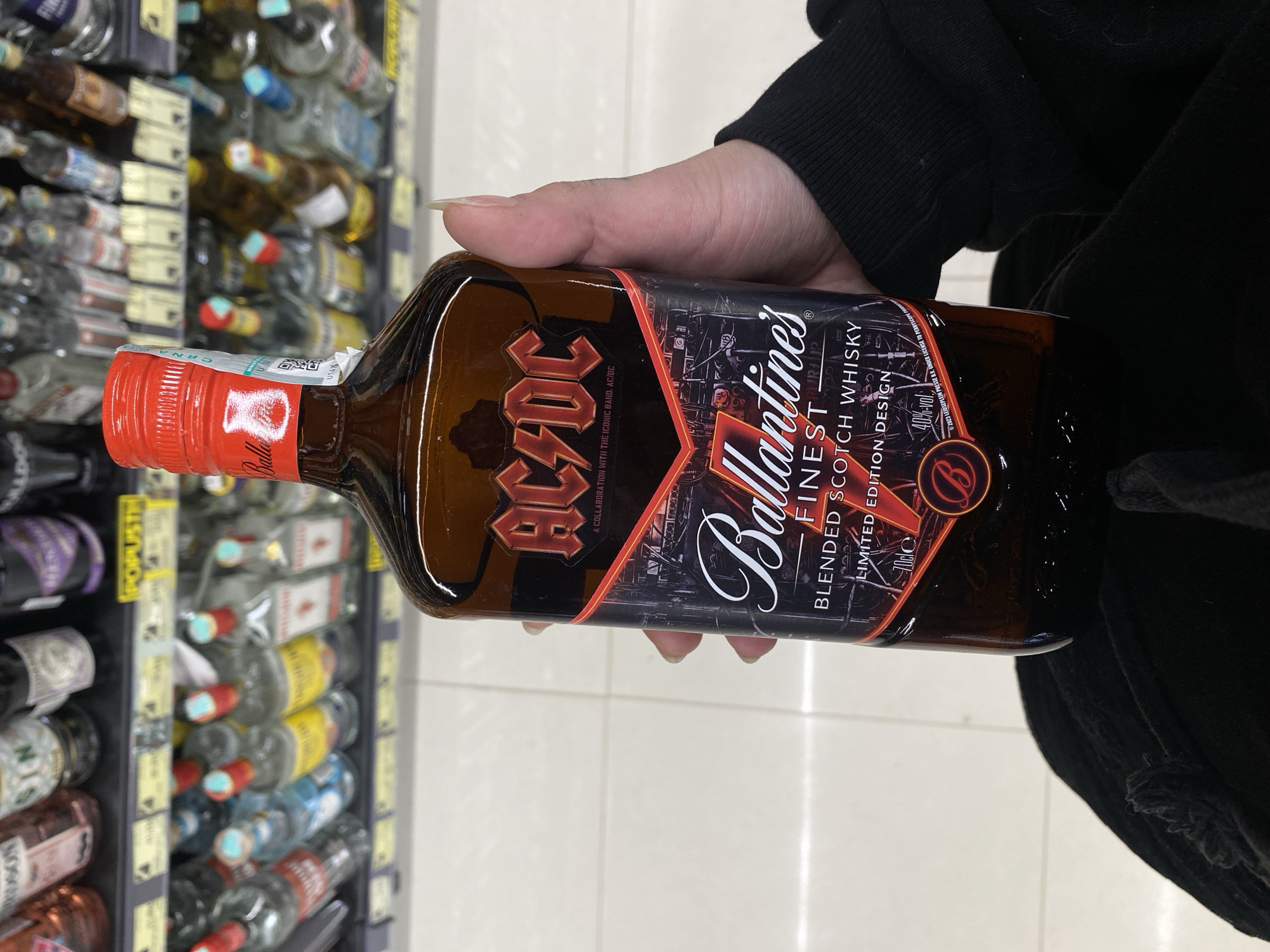 A person's hand is prominently featured, holding a dark brown glass bottle of whisky. The hand, with long, light-colored fingernails, is wearing a black long-sleeved top with ribbed cuffs. The bottle has a bright red screw cap, below which a label partially shows the brand "Ballantine's" and features a QR code and text including "CENA" and "CRNA U147".
The main label on the bottle is for "Ballantine's FINEST BLENDED SCOTCH WHISKY LIMITED EDITION DESIGN". It is a collaboration with the iconic band AC/DC, whose red lightning bolt logo is boldly displayed on the left side of the label, accompanied by the text "A COLLABORATION WITH THE ICONIC BAND. AC/DC". The label also specifies "70cl" and "40%-vol", along with copyright information from "2023". The background of the label features a dark, stylized cityscape.
In the background, out of focus, are rows of shelves stocked with various other bottles, indicating a retail store setting, likely an alcohol aisle. A yellow shelf label in the background is partially visible, displaying the word "POPUSTI," which translates to "discounts," further confirming the commercial environment. The floor appears to be a light-colored tiled surface. The scene takes place in a store, consistent with a retail location in Ulcinj, Montenegro.
