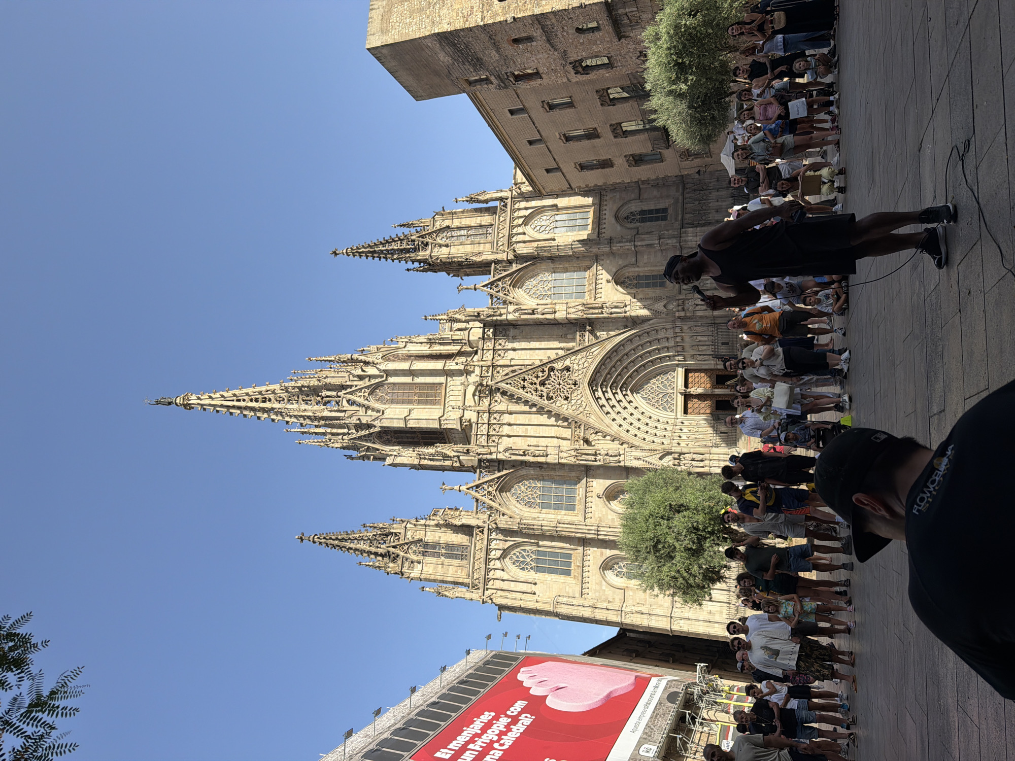 The image captures a street performer, a Black man in a black tank top and shorts, singing into a microphone on a cobblestone plaza in front of the Barcelona Cathedral in Barcelona, Spain. He is standing in the center of the frame, silhouetted against a large crowd gathered to watch him. The cathedral, a magnificent Gothic structure, dominates the background with its intricate facade and tall spires.
To the left of the cathedral, a large red billboard with white text and an illustration of a pink foot is visible. The plaza is populated by a diverse group of people of various ages and ethnicities, dressed in casual summer clothing. Some are standing, while others are seated on the ground. The overall atmosphere appears lively and public, with the performance drawing a significant audience. The sky is clear and blue, indicating good weather. In the foreground, the back of a person wearing a black baseball cap and a black t-shirt is visible, looking towards the performer.