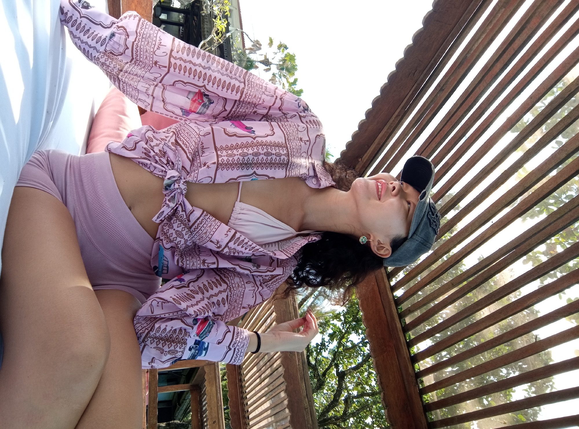 A woman is captured in a brightly lit, outdoor shot, reclining on a white surface with a pink cushion, looking upwards with a relaxed and happy expression. Her head is tilted back, revealing a green stud earring and a dark blue or grey baseball cap worn backward.
She is dressed in a light pink bikini top, light pink ribbed athletic shorts, and a sheer, oversized pink cover-up with a brown patterned design. The cover-up is tied loosely at her waist. The fabric pattern features various motifs including leaves, geometric shapes, and stylized illustrations of classic cars or vans and palm trees. Notably, the words "bali" and "Indonesia" are visibly integrated into the fabric's design, alongside other stylized text fragments such as "chup" and "lol." A black hair tie or bracelet is on her left wrist.
The setting is under a slatted wooden roof or pergola, allowing sunlight to stream through. Lush green foliage and trees are visible in the background, suggesting a tropical environment. The bright lighting indicates it is daytime with clear weather. The overall scene conveys a mood of leisure and contentment, consistent with a relaxed moment in North Kuta, Indonesia.