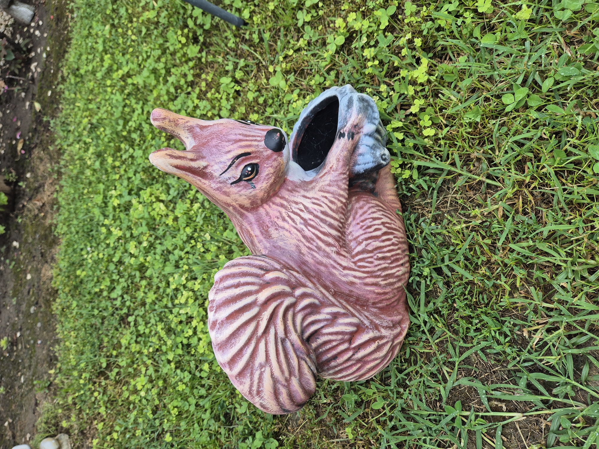 An artistic sculpture of a pink squirrel, with stripes of cream and darker pink, sits on a patch of green grass. The squirrel is in a seated position, with its tail curled up over its back. It is holding a gray, hollowed-out object in its paws, resembling a nut or a small bowl. The squirrel has black eyes with visible eyelashes and a black nose. The background consists of more grass and some dirt. The image is taken from a slightly elevated angle, looking down at the sculpture.