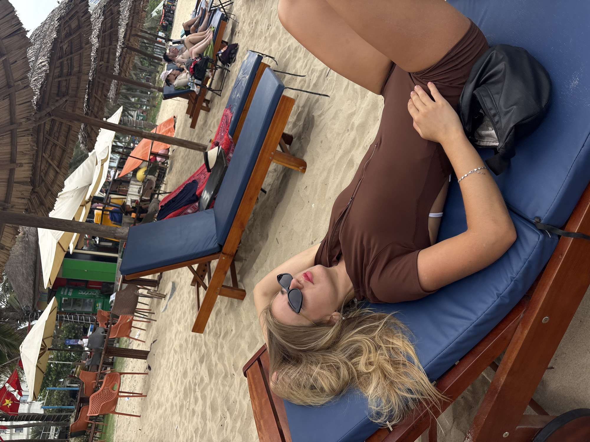 The image, oriented on its side, depicts a young woman relaxing on a beach, likely in Đà Nẵng, Vietnam. She is lying on her back on a blue-cushioned wooden lounge chair situated directly on the sand. Her long, wavy blonde hair flows out behind her head, and she wears dark sunglasses. Dressed in a brown, long-sleeved one-piece swimsuit or bodysuit, she also has a silver chain bracelet on her left wrist. A black bag rests on the chair next to her.
The setting is a vibrant beach scene, with numerous other blue-cushioned lounge chairs visible in the midground and background. Many of these chairs are placed under traditional thatched palm-leaf canopies, providing ample shade. Other, more modern white and orange umbrellas are also scattered across the sandy expanse. Several other people are visible in the distance, similarly relaxing on lounge chairs.
Further back, there are indications of beachside establishments, including tables with orange plastic chairs and a green structure, suggesting cafes or service areas. Palm trees are interspersed throughout the scene, enhancing the tropical atmosphere. A significant detail confirming the location is the national flag of Vietnam, a red flag with a yellow star, visible towards the bottom left of the wider shot.
The scene takes place during the daytime, with soft lighting suggesting either an overcast sky or the effective shade provided by the canopies. The overall mood is one of leisure and tranquility, capturing a typical relaxing day at a beach resort.