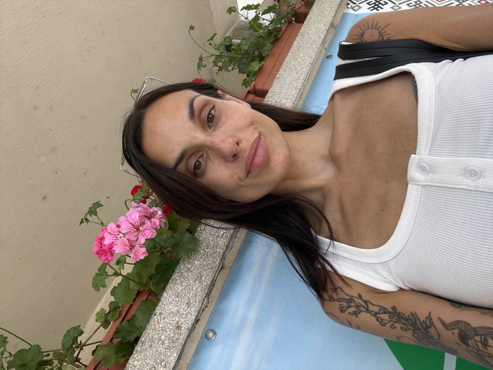In Pordim, Bulgaria, a selfie of a woman with long dark hair is captured outdoors. She is wearing a white ribbed tank top. A planter box with pink flowers and green leaves is positioned behind her. She has several tattoos on her left arm, including one of a bird, and a sun tattoo on her right shoulder. The background features a light-colored building facade and a blue wall with a sky pattern. The overall scene suggests a casual outdoor setting.