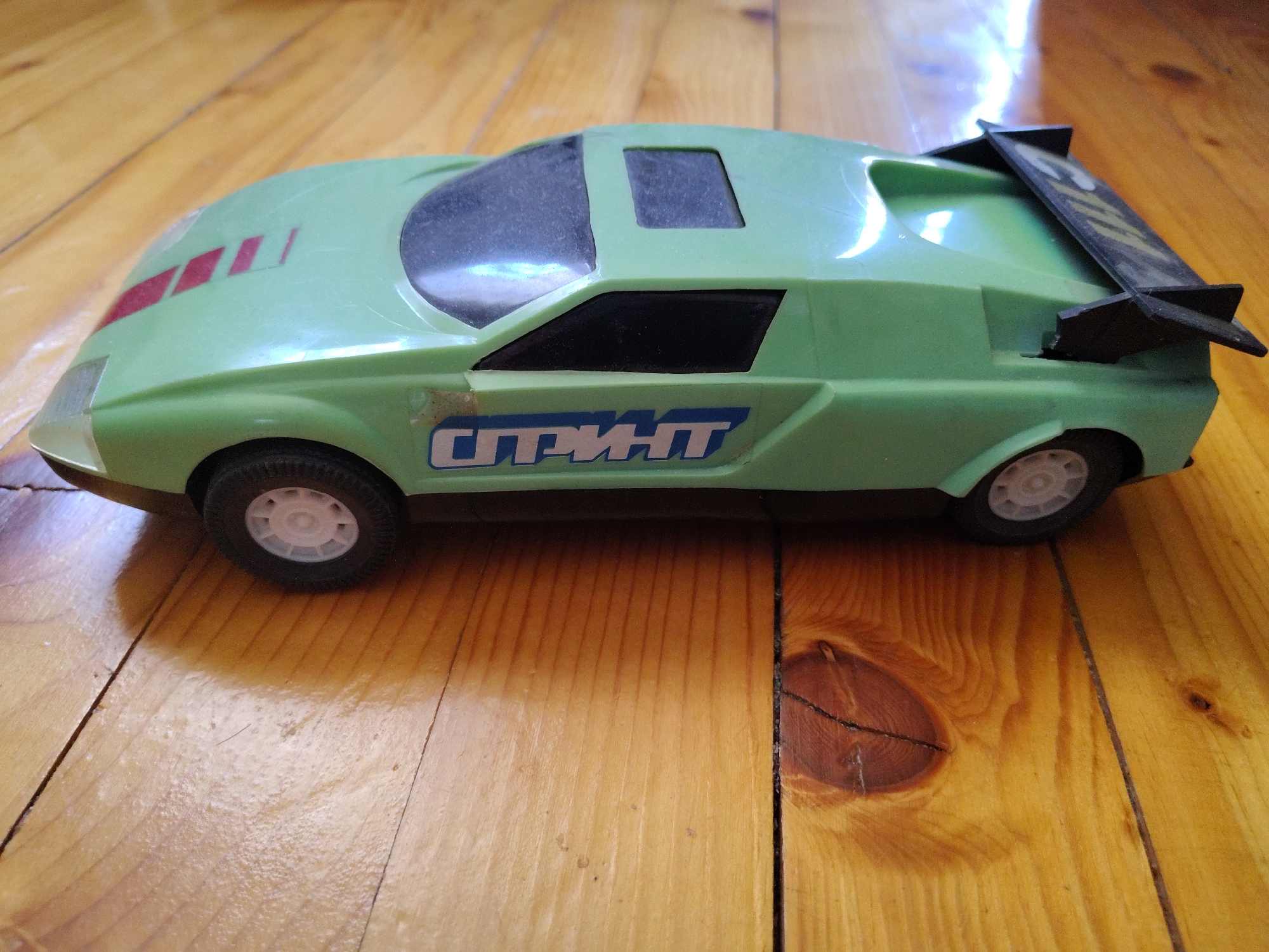 A green toy car with a spoiler and red stripes on the hood is positioned on a wooden floor. The car has the word "СПРИНТ" (SPRINT) written on its side in blue letters. The floor appears to be made of light-colored wooden planks. The lighting suggests it is indoors, with natural light entering from an unseen source, creating a bright area on the floor to the upper right. The scene does not depict any specific location in Kyustendil, Bulgaria, or any people.