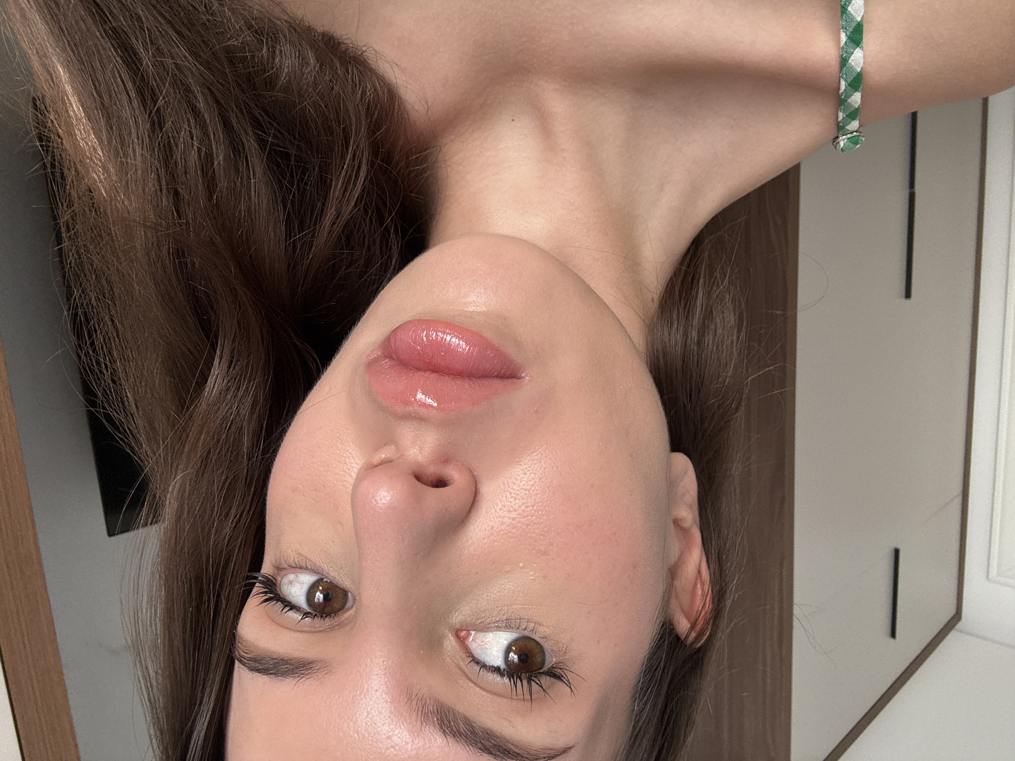 A close-up, medium shot captures a young woman's face and upper torso from a slightly low angle. She has medium-brown hair that is parted and falls around her face and shoulders. Her eyes, with brown irises and defined eyelashes, gaze to her left, while her nose is angled slightly upwards. Her lips are full and coated in a glossy pink lip product, reflecting light. Her skin appears smooth and dewy. She is wearing a green and white checkered strap with a thin, silver ring on the strap over her right shoulder. The background consists of a wall with vertical dark lines, and to the right, a darker wooden surface. The lighting is bright and diffused. The image is likely taken indoors.