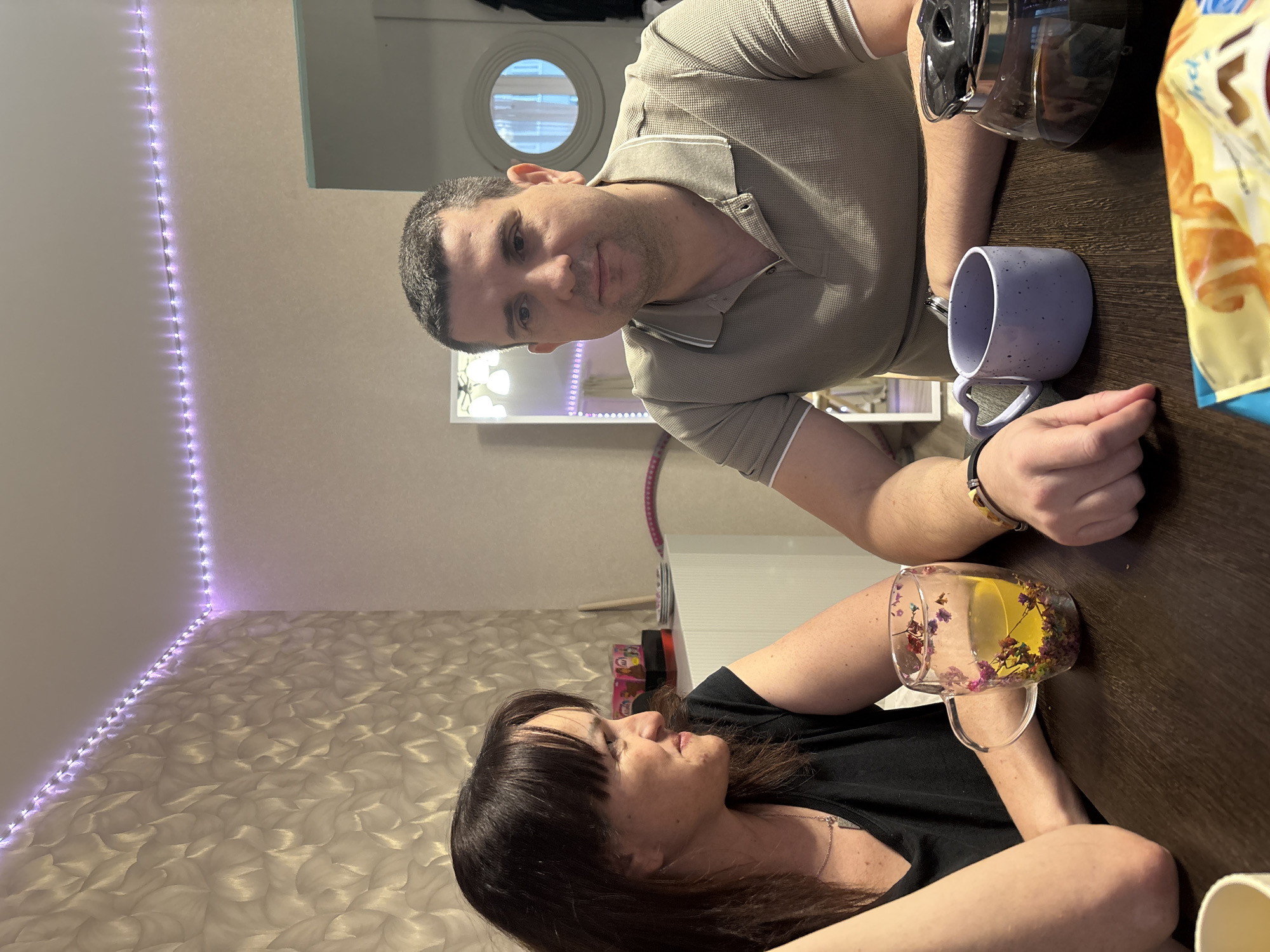 A man and woman are seated at a table indoors. The man is fair-skinned, with short, dark hair, wearing a collared shirt, and looking at the camera. He has a mug and a coffee pot near him on the table. The woman has dark hair and is wearing a black shirt. She is looking at the man and has a clear mug of tea with flowers in it in front of her. The table is dark brown. There is a wall with purple LED lights along the corner. The location is Кондратово, Russia.