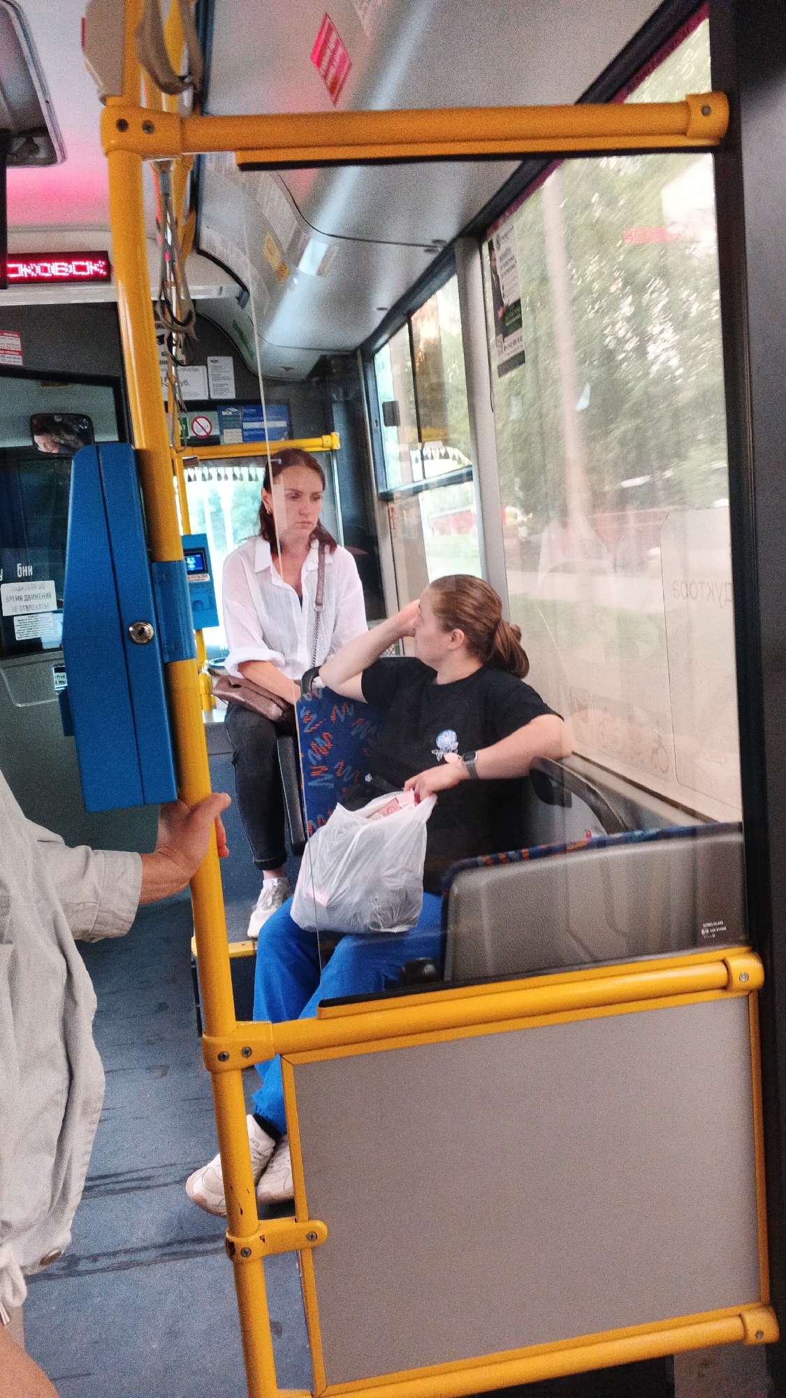The image depicts the interior of a public bus in Izhevsk, Russia. Two women are seated. The woman in the foreground on the right is wearing a black t-shirt and blue pants, with her hair tied back in a ponytail. She is resting her head on her hand and holding a white plastic bag. The woman behind her on the left is wearing a white shirt and dark pants, with her hair down. She appears to be looking forward. The bus interior features yellow and blue handrails and grey seats with a patterned fabric. A sign with Cyrillic text is visible in the background. The environment suggests an urban setting, likely during daylight hours.