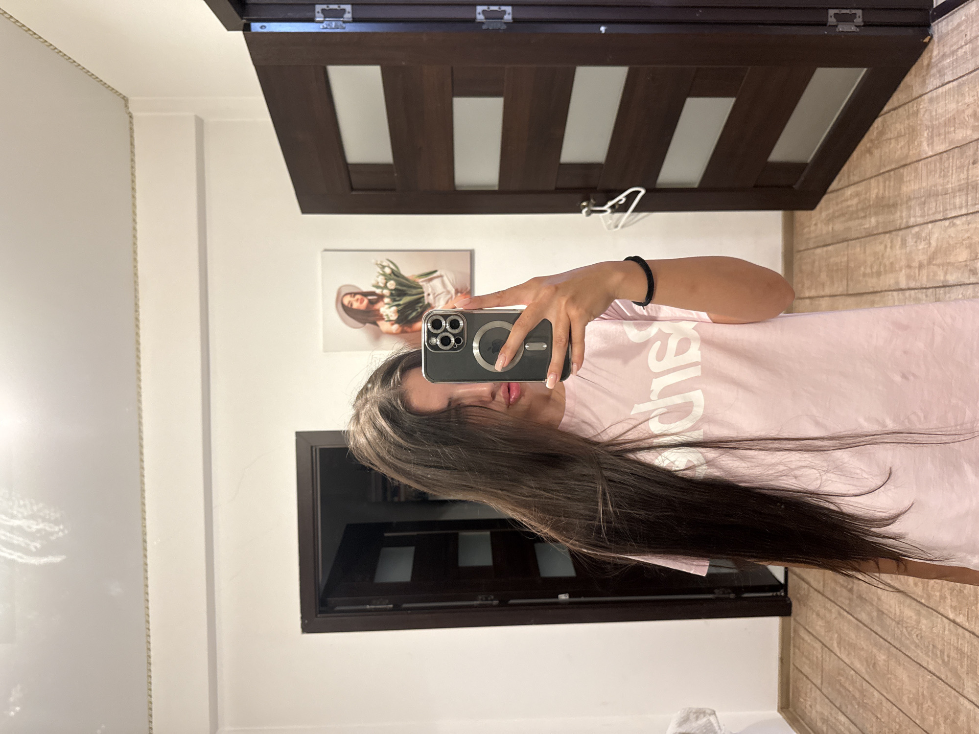 A young woman with long, dark brown hair is taking a selfie in a hallway. She is wearing a light pink t-shirt with white lettering. Her left hand is holding a black smartphone with a silver camera module, and her right hand is extended, obscuring part of her face. She has a black hair tie on her right wrist. In the background, there is a dark wood door on the right, with a white hanger hanging from it. To the left of that door, there is a framed picture on the wall depicting a woman in a white hat holding a bouquet of white tulips. Another dark wood door is visible on the left side of the hallway. The floor is made of light-colored wooden planks. The walls are white, and a decorative gold trim runs along the top of the wall. This scene is likely taking place in a residential apartment or hallway, common in urban environments such as Chișinău, Moldova.