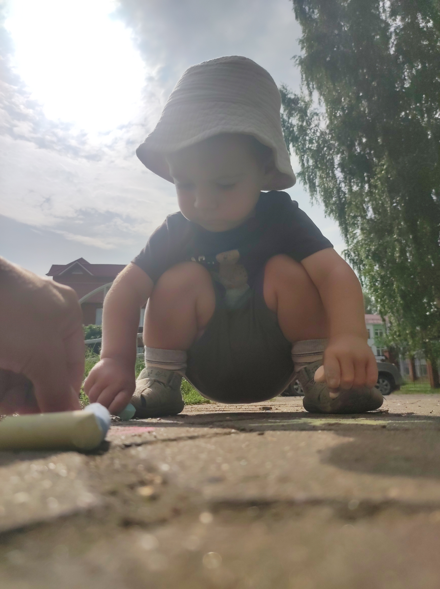 An outdoor, low-angle, full shot shows a young child squatting on a paved surface. The child, wearing a bucket hat, dark t-shirt, shorts, socks, and light-colored shoes, is looking down at chalk in their hands. To the left of the child, a hand is visible, reaching towards the chalk. A piece of yellow chalk lies on the ground in the foreground. The background includes a building with a red roof, a green tree, and a car. The sky is partly cloudy and bright, suggesting daytime. The scene appears to be in an urban or suburban environment, possibly a park or residential area.