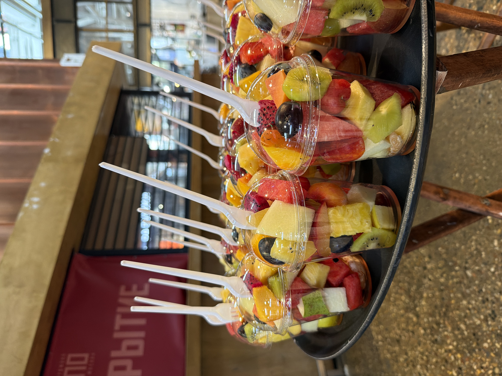 The image shows a tray of plastic cups filled with various cut fruits, each cup containing a white plastic fork. The fruits appear to include watermelon, kiwi, grapes, pineapple, dragon fruit, oranges, and possibly others. The cups are stacked tightly on a black tray. The background suggests an indoor location, with a sign in Russian script and a partially visible staircase. Based on the Cyrillic text and details of the environment, the location is likely in Moscow, Russia.