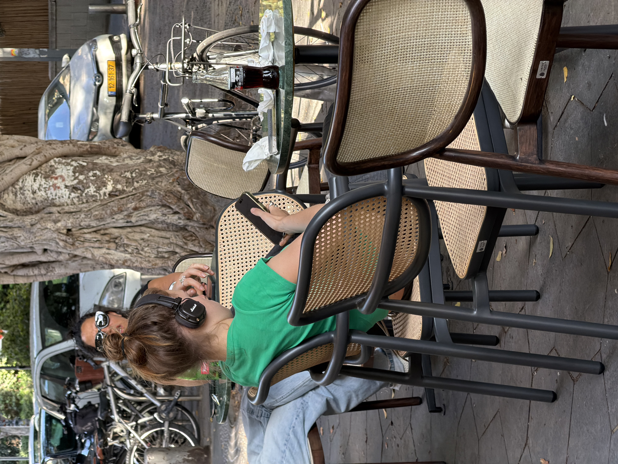 This image depicts an outdoor cafe scene in Tel Aviv, Israel. Two women are seated at tables on a dark gray flagstone pavement. In the foreground, a woman with light brown hair tied in a bun is seen from behind, wearing a bright green t-shirt and black Marshall over-ear headphones. She holds a black smartphone with a gold-colored bezel in her left hand, looking at the screen. Her lower body, clad in light blue jeans, is visible to the left. She is seated on a dark gray chair with a woven cane back and seat.
Behind and to the right of the foreground woman, a second woman is partially visible, facing the viewer. She wears dark sunglasses and a dark top, with dark, wavy hair. Her right hand, adorned with a silver ring, rests on the table. She is also seated on a chair with a cane back. Between the two women, a round green marble-topped table holds a glass Coca-Cola bottle, crumpled white napkins, and a metallic object.
A large tree with light-colored, rough bark stands prominently on the right side of the image, partially obscuring the background. In the background, several bicycles are parked to the left of the tree, and another bicycle with a front basket is visible behind the green table. A white Toyota sedan with an Israeli license plate "12-161-37" is parked on the street in the middle-right background. Another white car is partially visible further to the left. Green foliage is present in the upper left background, and a light brown wooden slatted wall or fence is visible in the far background on the right. The scene is brightly lit by daylight. One chair in the far right foreground has a dark wooden frame with a cane seat, featuring a "UNITED SEATS" label.